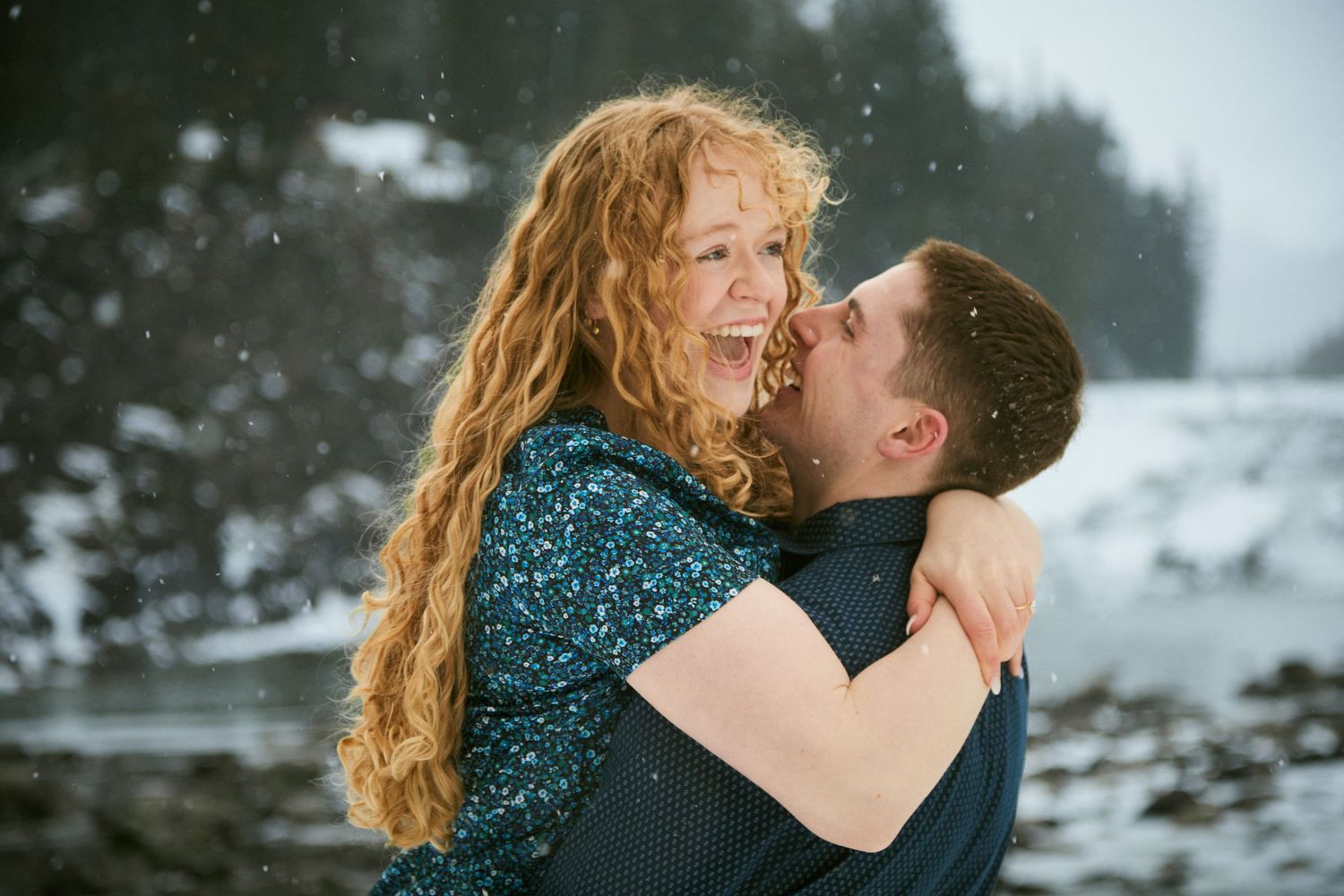 Couple embracing and laughing in snowy winter landscape, woman with red curly hair wearing teal sequin dress.