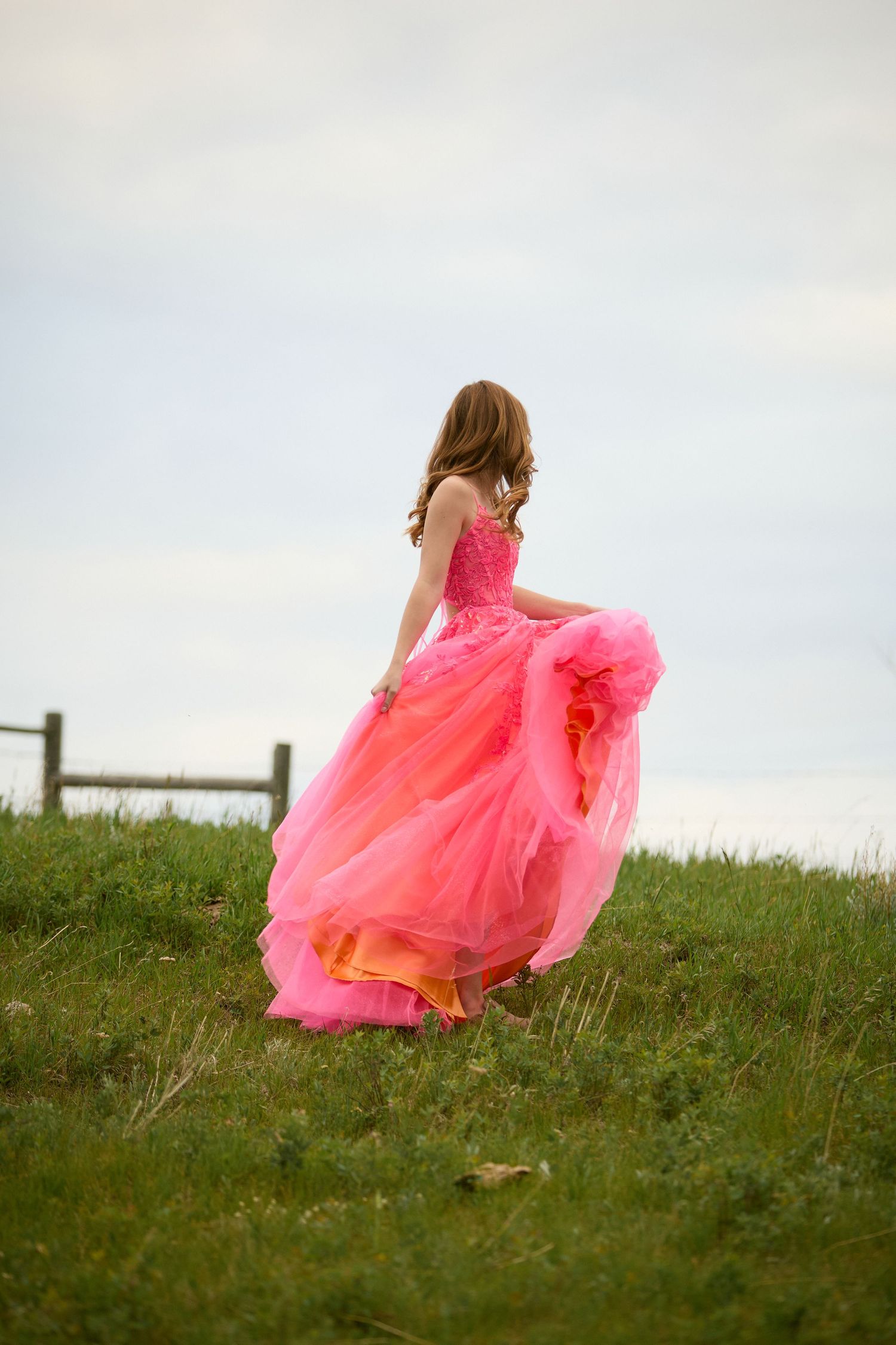 Young woman in flowing pink ballgown walks through green meadow, dress billowing in wind near rustic wooden fence.