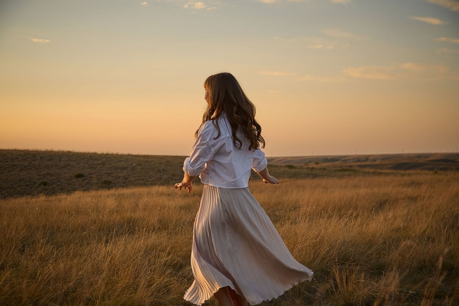 A person in a flowing white dress twirls in a golden field at sunset.