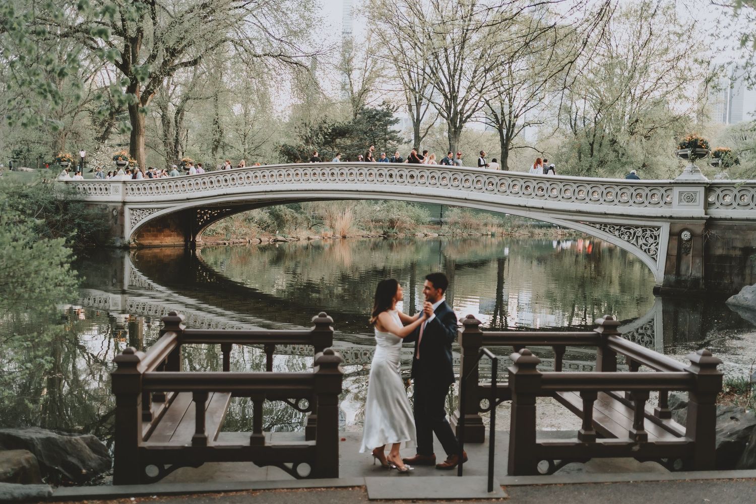 A romantic couple poses under a stone bridge in a scenic park setting during their wedding photoshoot.