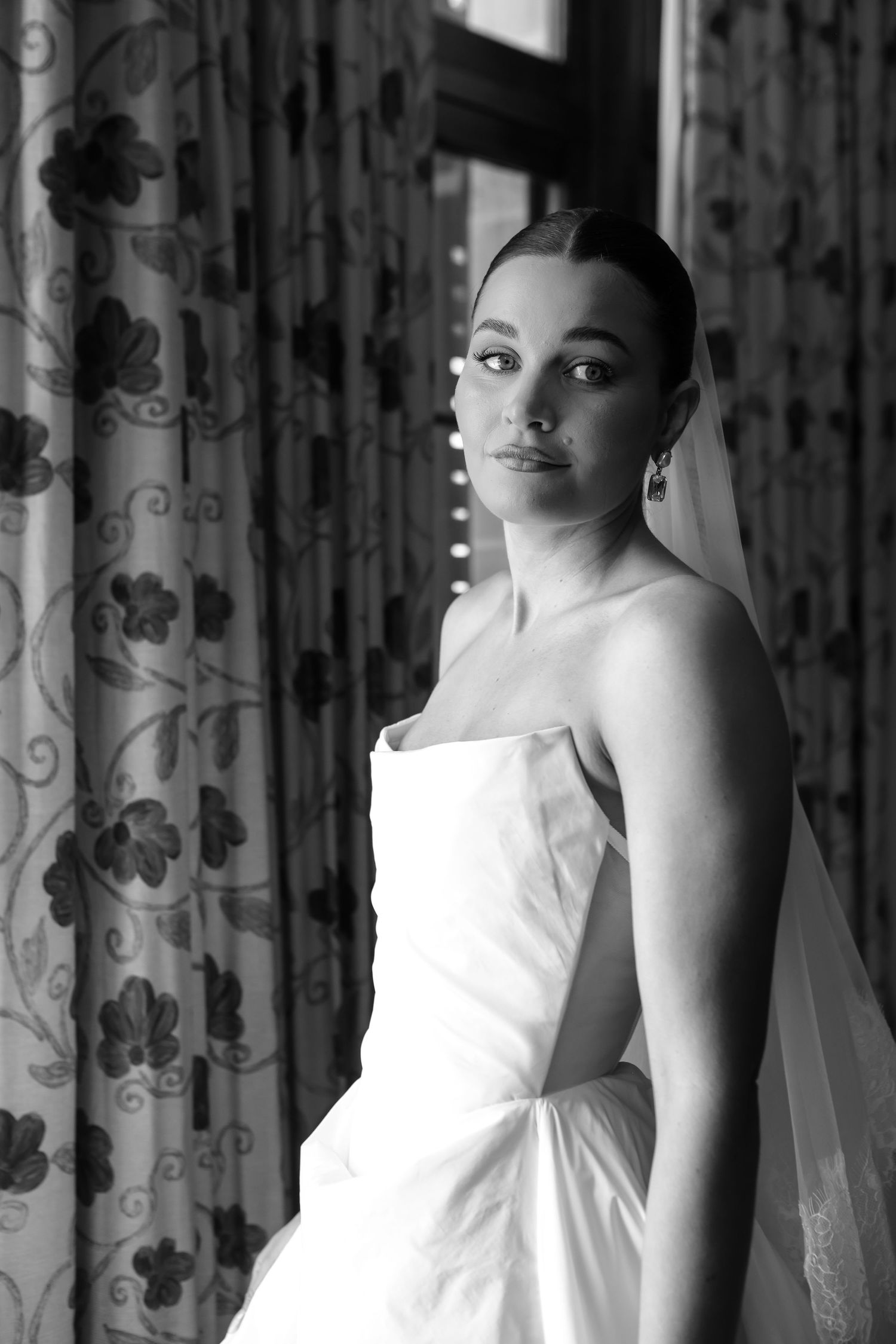 A black and white portrait of a bride in a sleeveless wedding dress standing against patterned curtains.