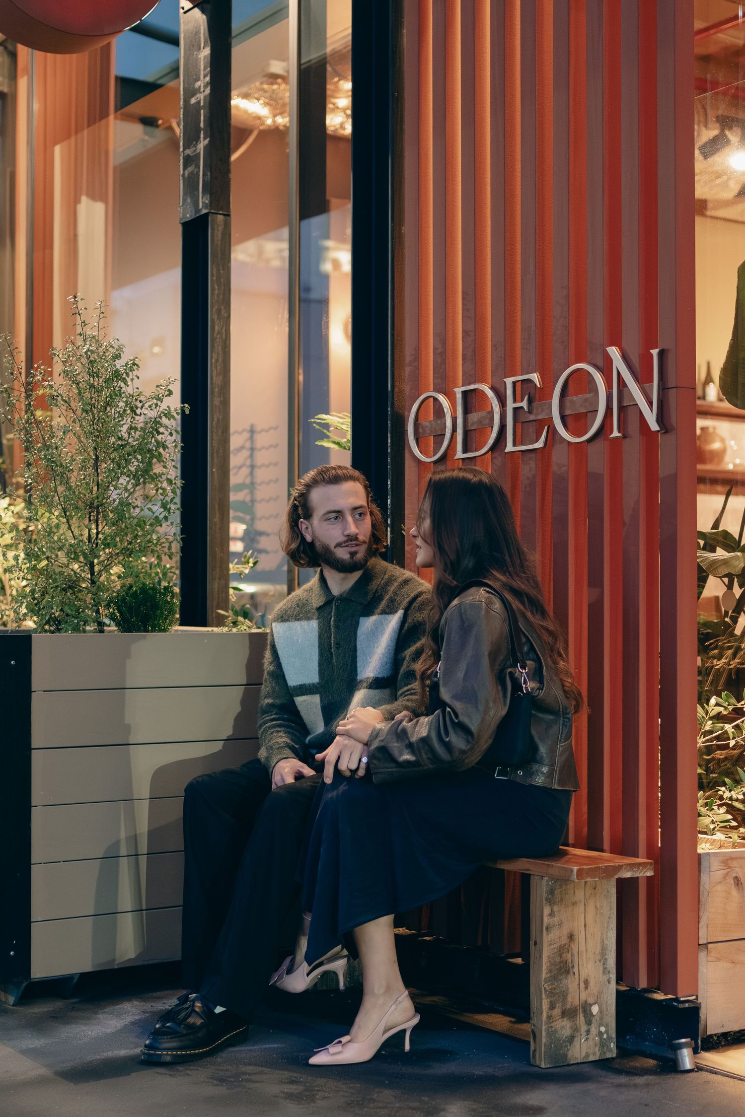 Couple sitting on wooden bench outside Odeon building having coffee together.