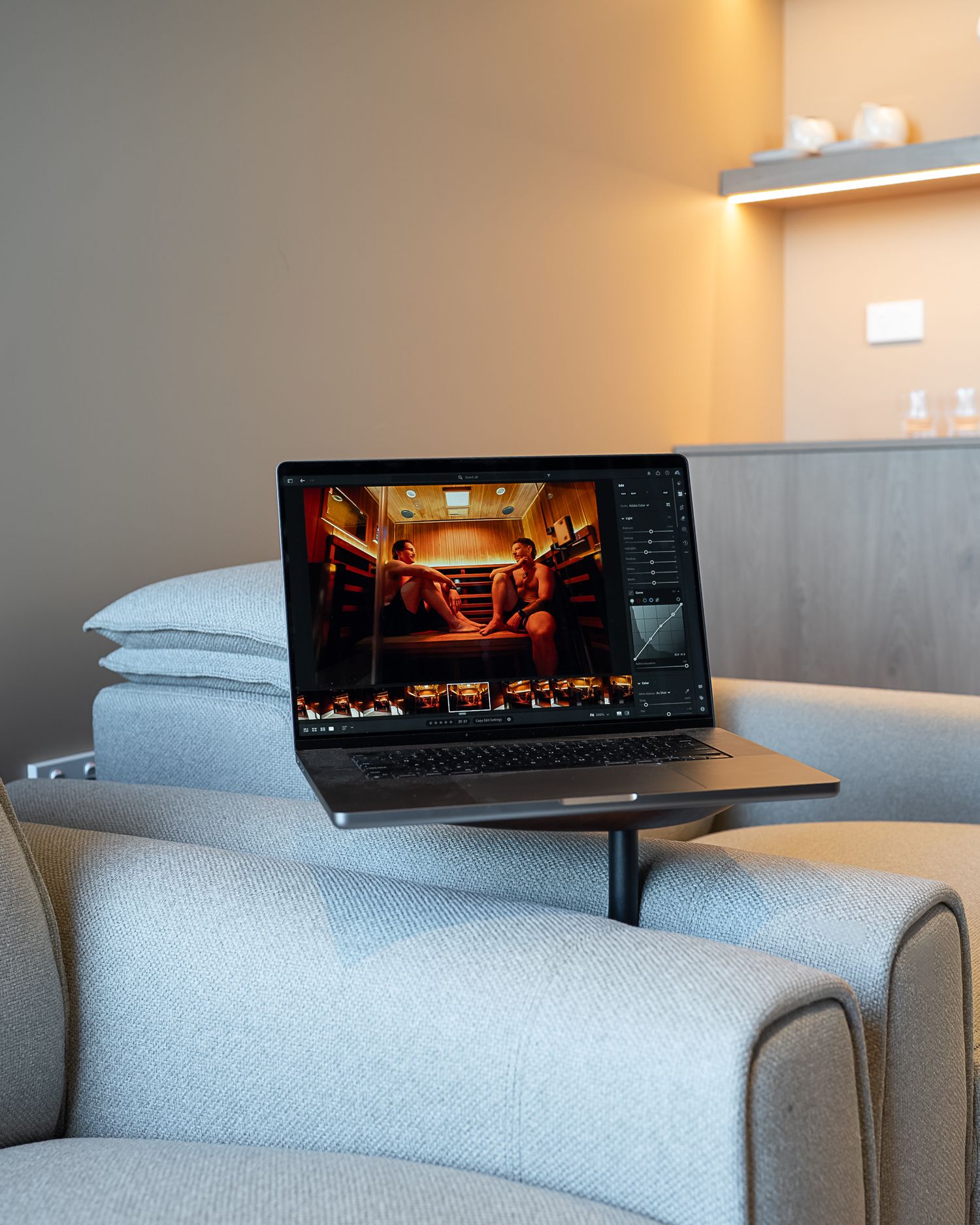 A laptop displaying photo editing software sits on a side table beside a cozy gray armchair in a modern room.