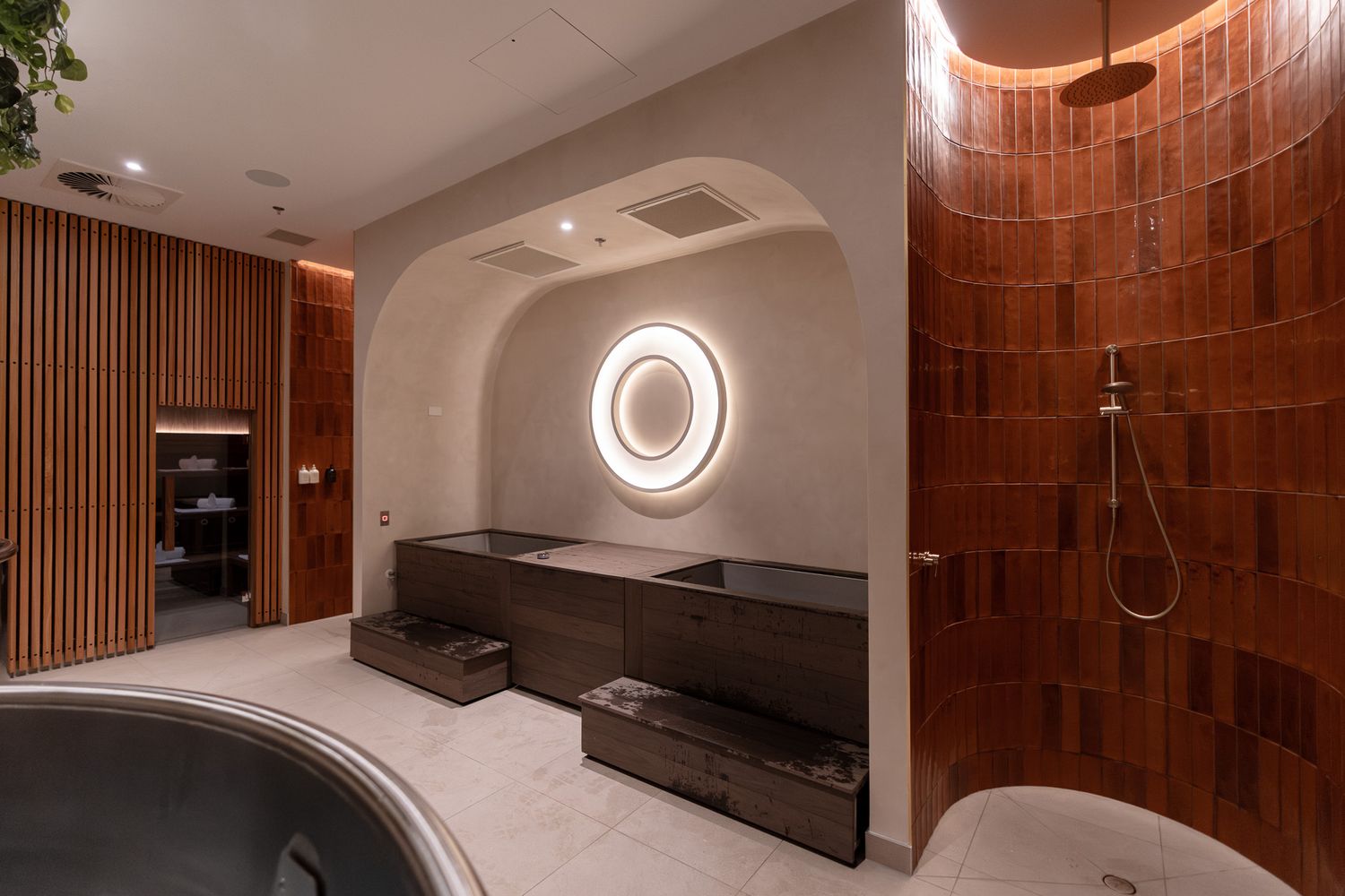 Luxurious spa bathroom featuring dark soaking tubs, brick accent wall, rain shower, and arched alcove with circular mirror.