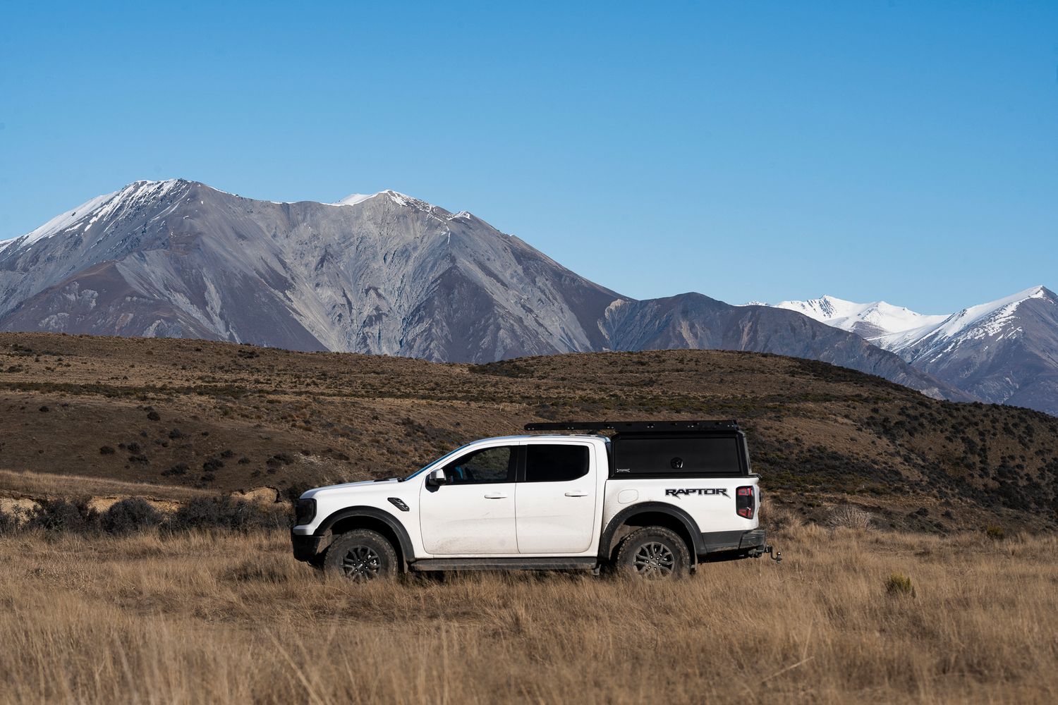 White pickup truck parked against a dramatic mountain landscape with snow-capped peaks.