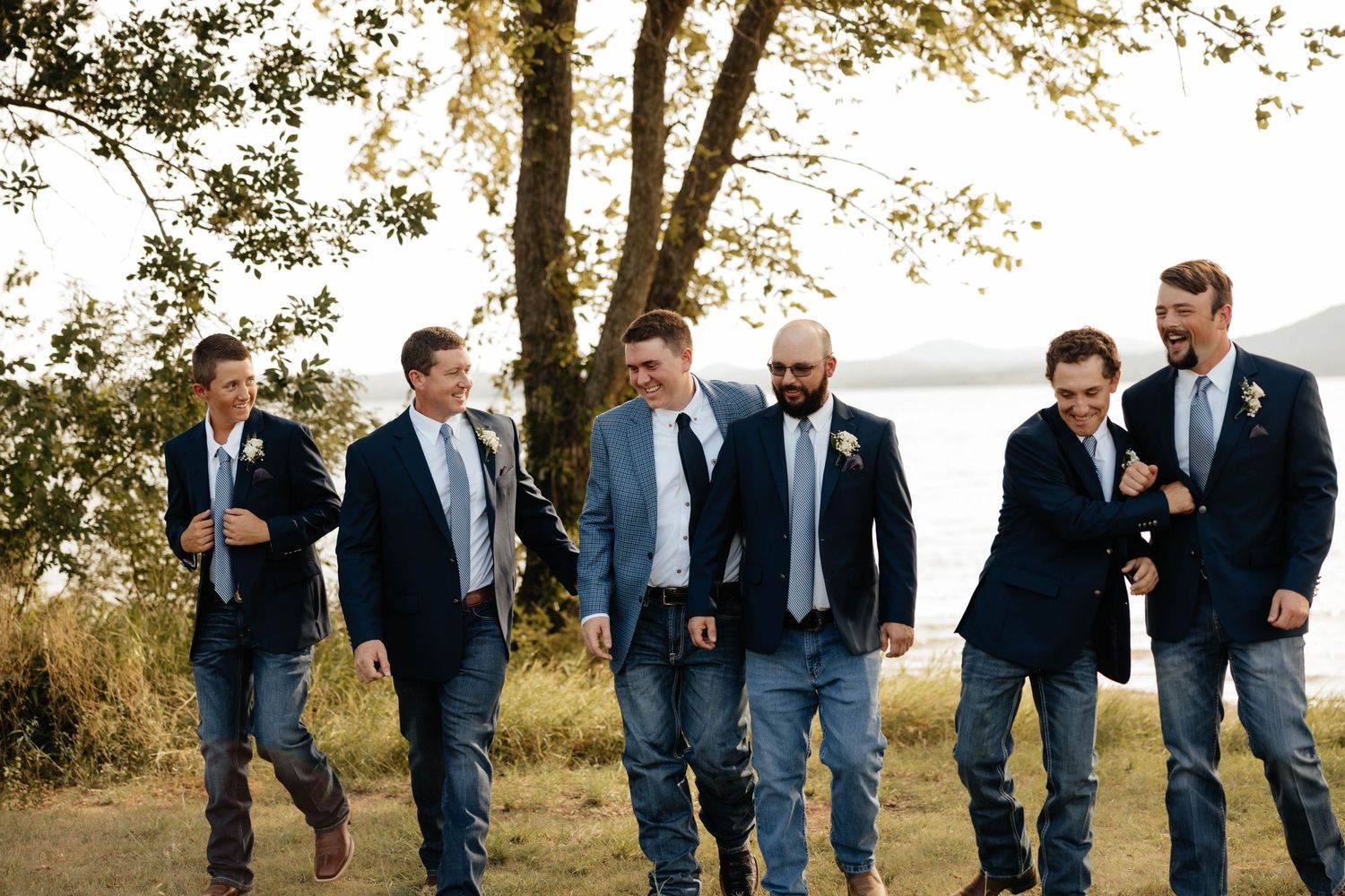 Groom and five groomsmen in navy blazers and jeans laugh while walking together near a lakeside tree.