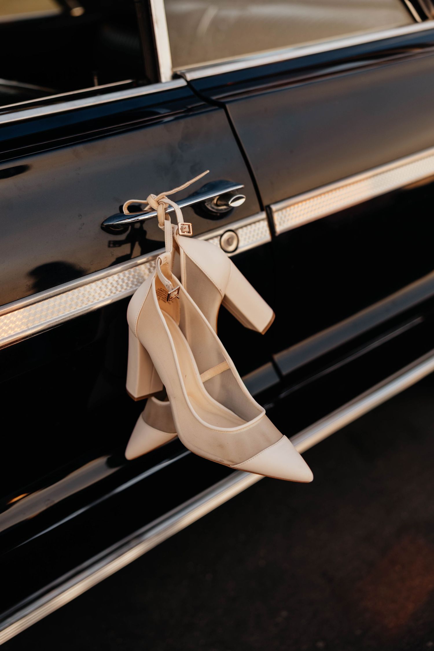Elegant cream block heel bridal shoes hanging from a vintage black car door handle.