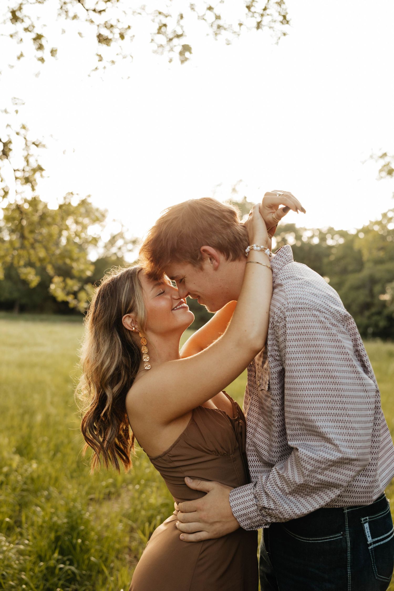 Couple shares a romantic near-kiss in golden sunlight in a lush green field during engagement session.