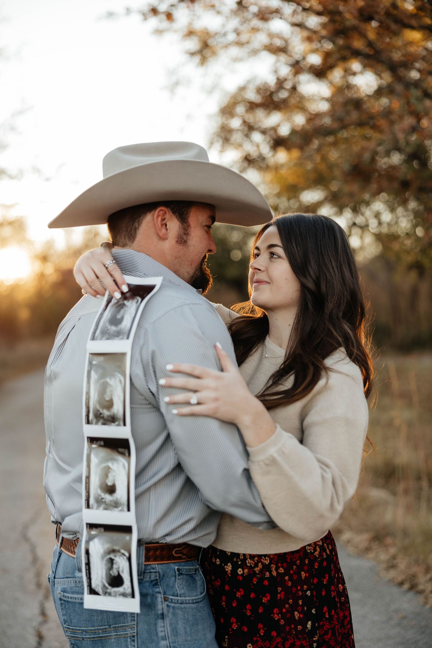 Couple embracing on dirt road at sunset, man in cowboy hat holding ultrasound photos behind back.