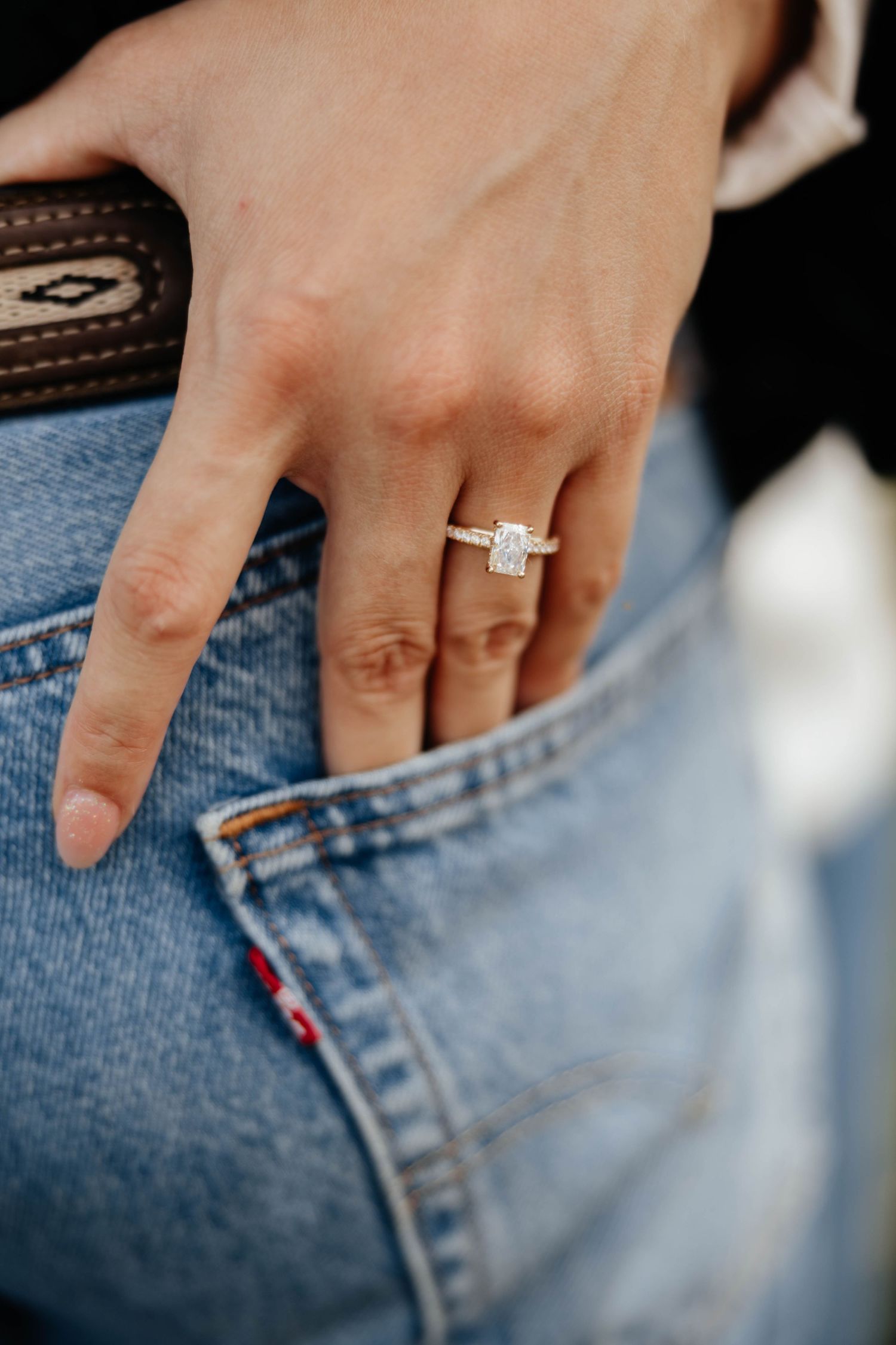 Close-up of womans hand wearing elegant emerald-cut diamond engagement ring on denim jeans pocket.