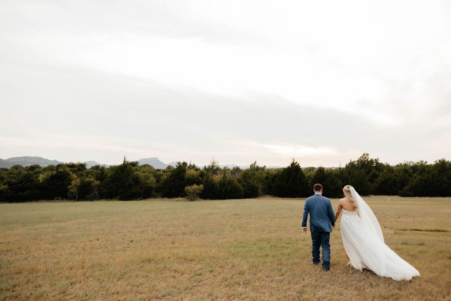 Bride and groom walking hand in hand through an open field with mountain views and trees at dusk.