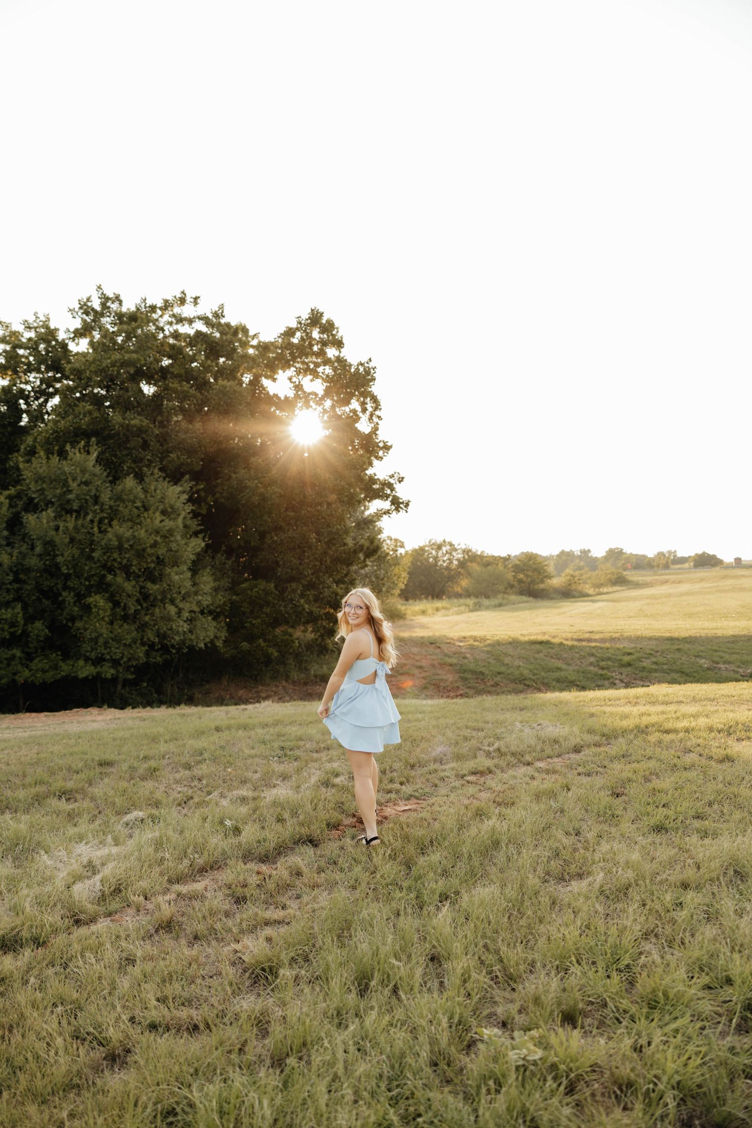 Young woman in light blue dress walking through green field at golden sunset hour.