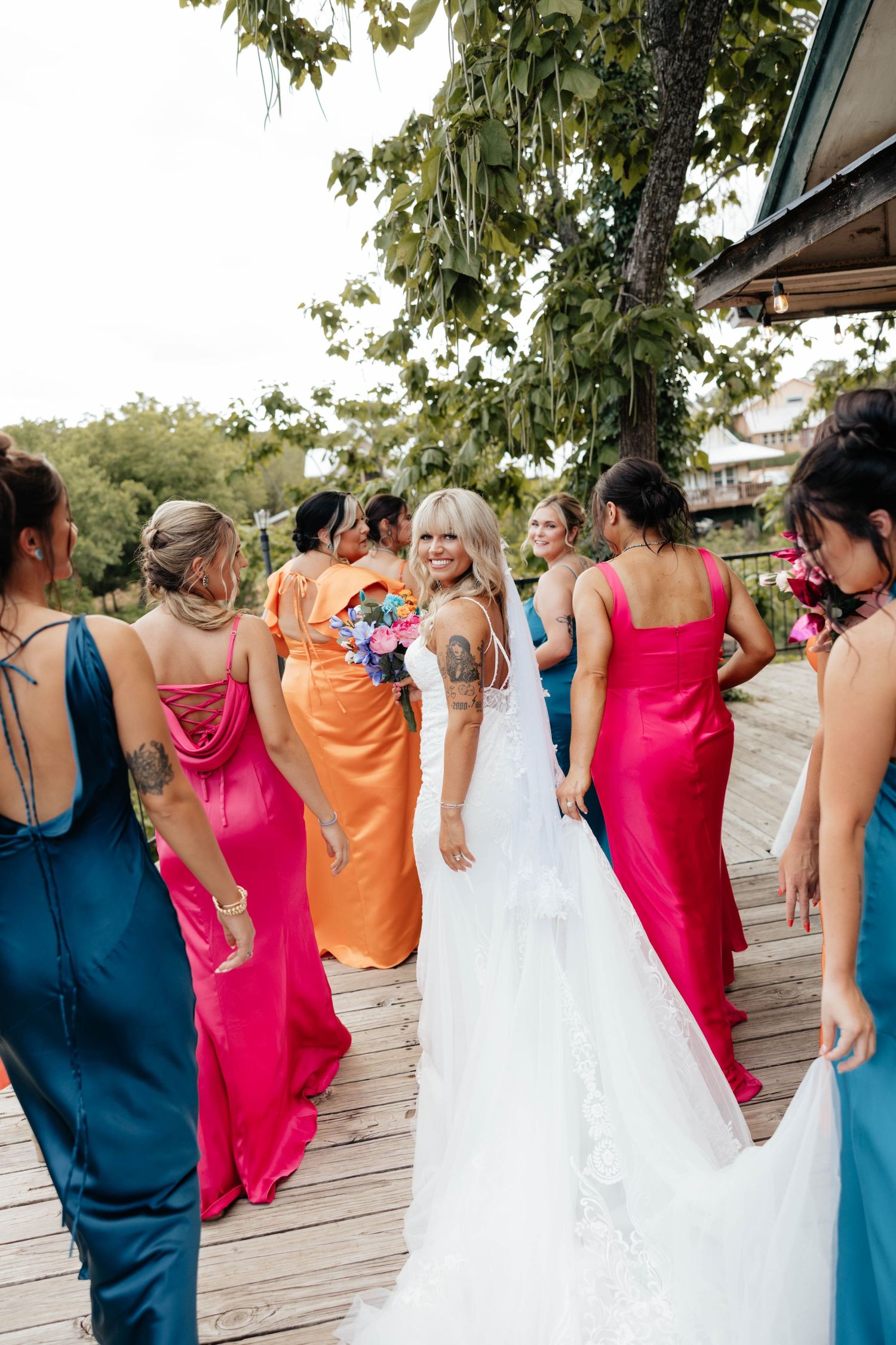 Bride in white gown turns to smile at camera while walking with colorful bridesmaids on a wooden deck.