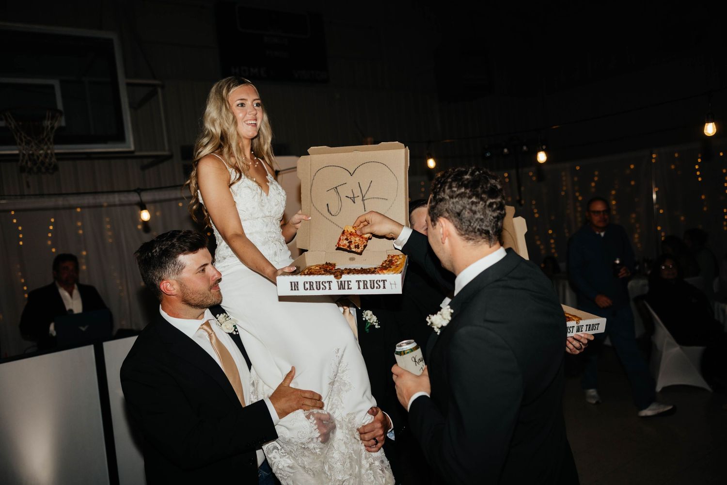 Bride seated on chair being lifted by groomsmen holding pizza at wedding reception.