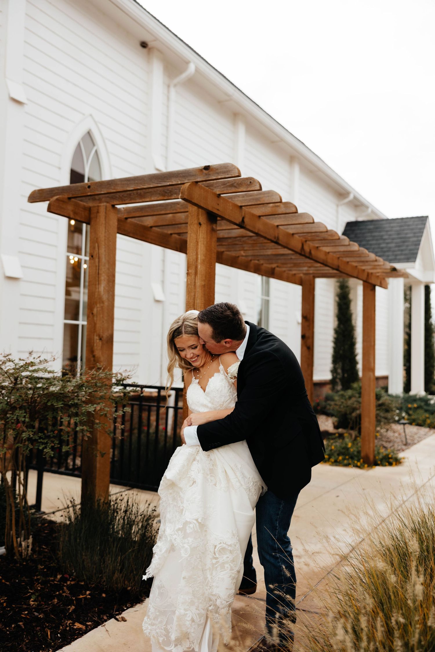 Groom kissing bride on cheek outside white wedding venue under wooden pergola after ceremony.