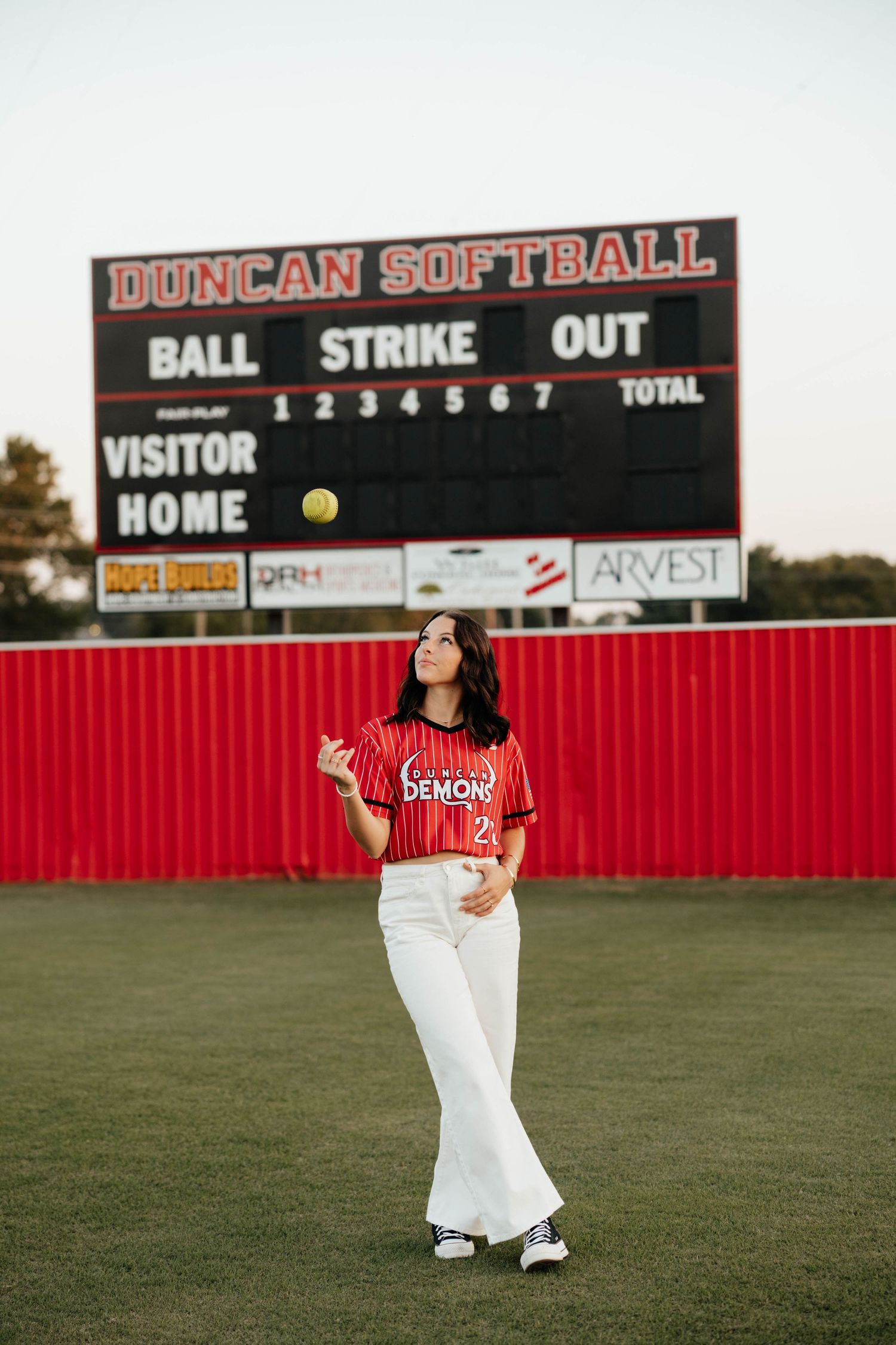 Duncan Softball player in red pinstripe jersey tosses ball near scoreboard at dusk.