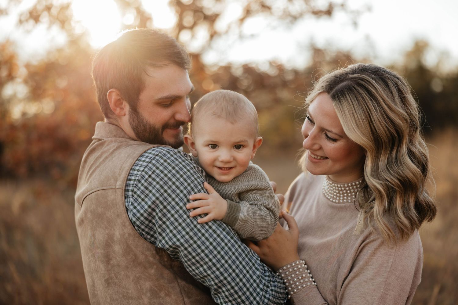 Happy family with parents holding their smiling baby boy in a golden autumn field at sunset.