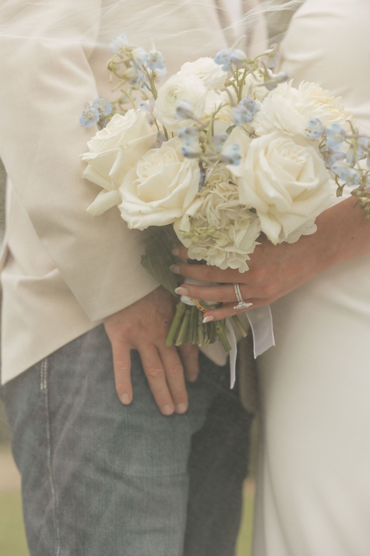 Bride holding white rose and blue flower wedding bouquet while groom places hand on her waist in casual attire.