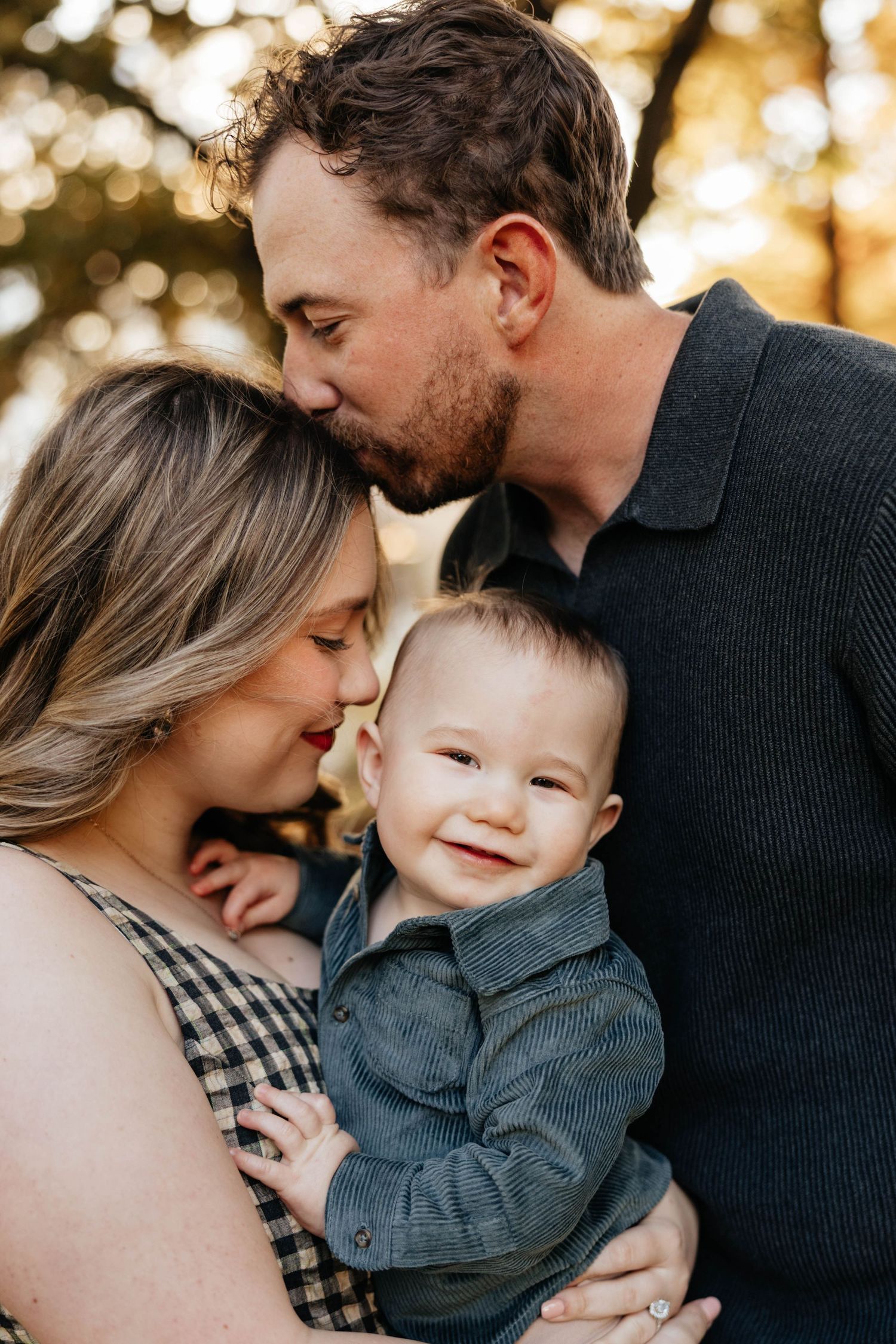 Parents kissing smiling baby boy in denim shirt outdoors at golden hour in family portrait.