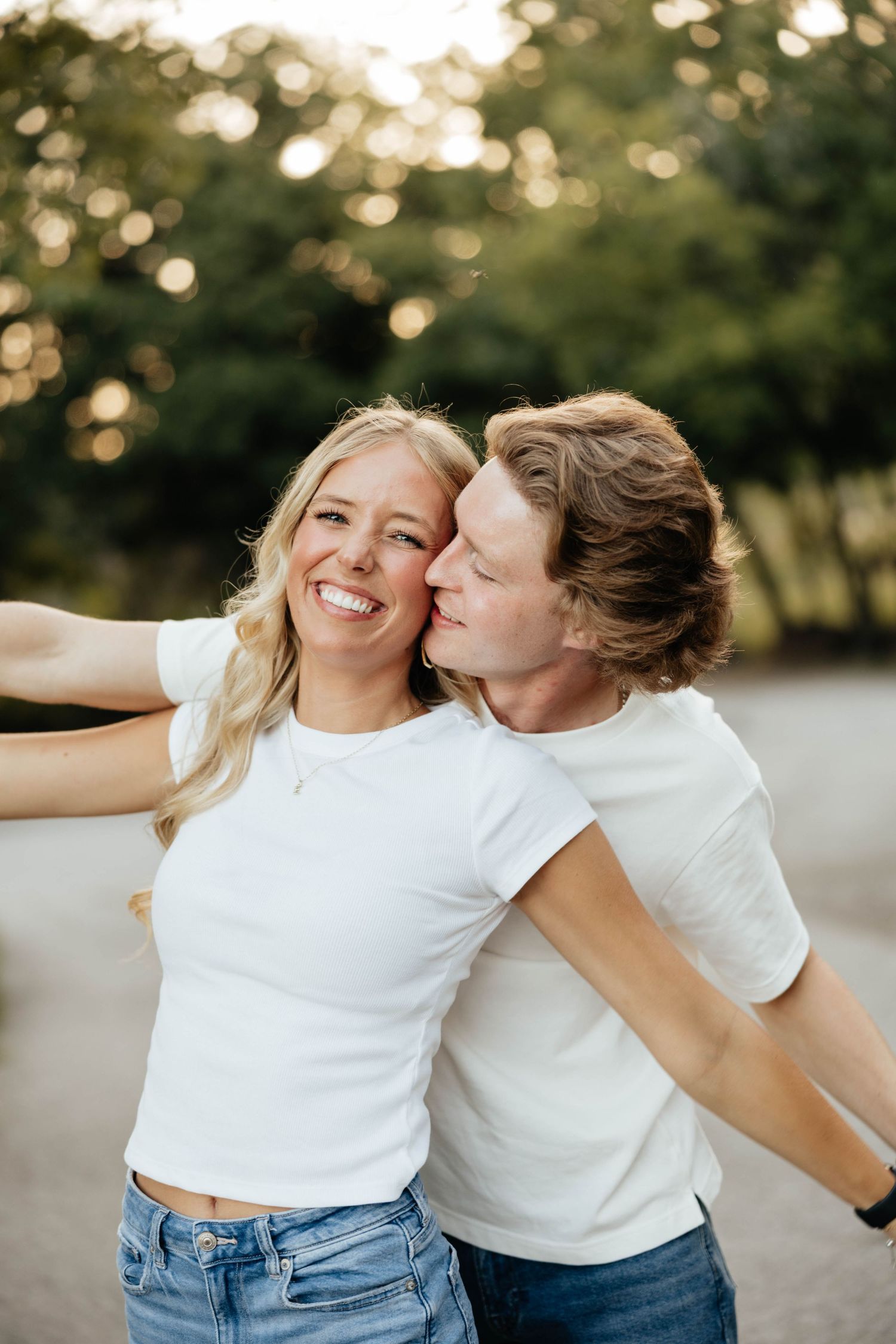 Smiling blonde woman in white crop top gets a cheek kiss from her partner outdoors near lush green trees.