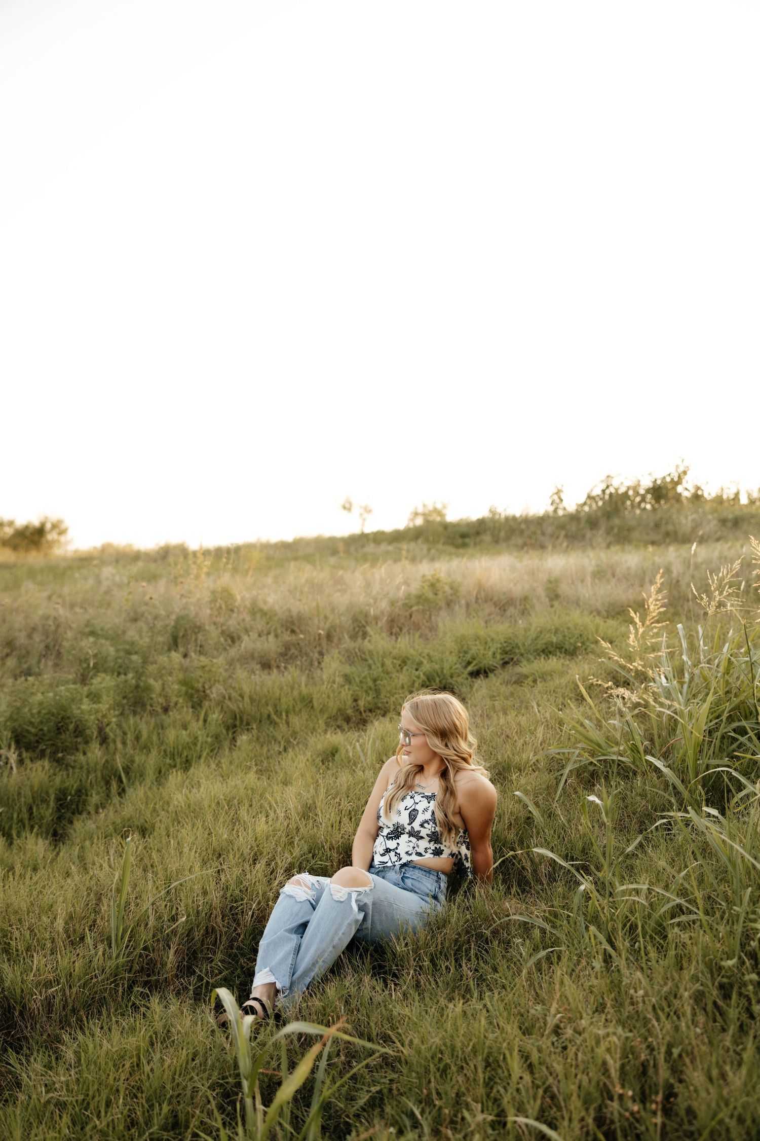 Blonde woman sitting in tall grass field at sunset wearing floral top and ripped jeans.