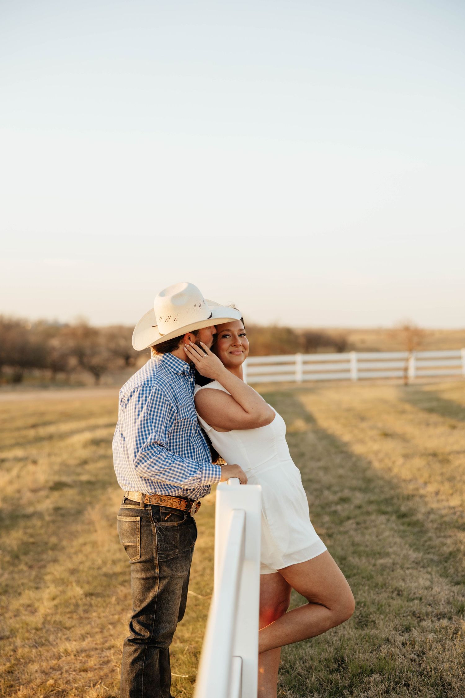 Couple in cowboy hats embracing by a white fence at golden hour on a ranch.