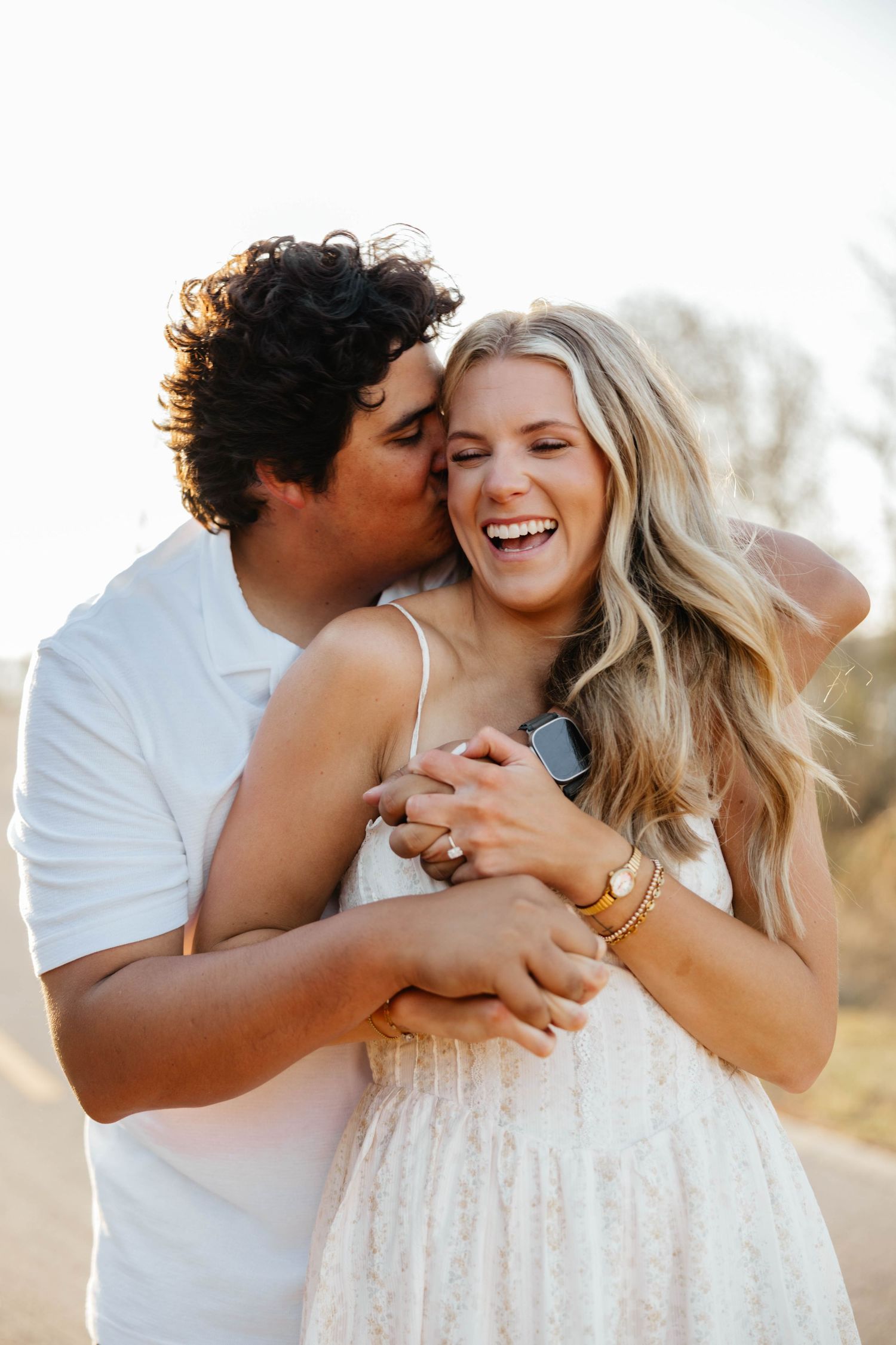 Man in white shirt kissing laughing blonde woman during sunny outdoor engagement photos.