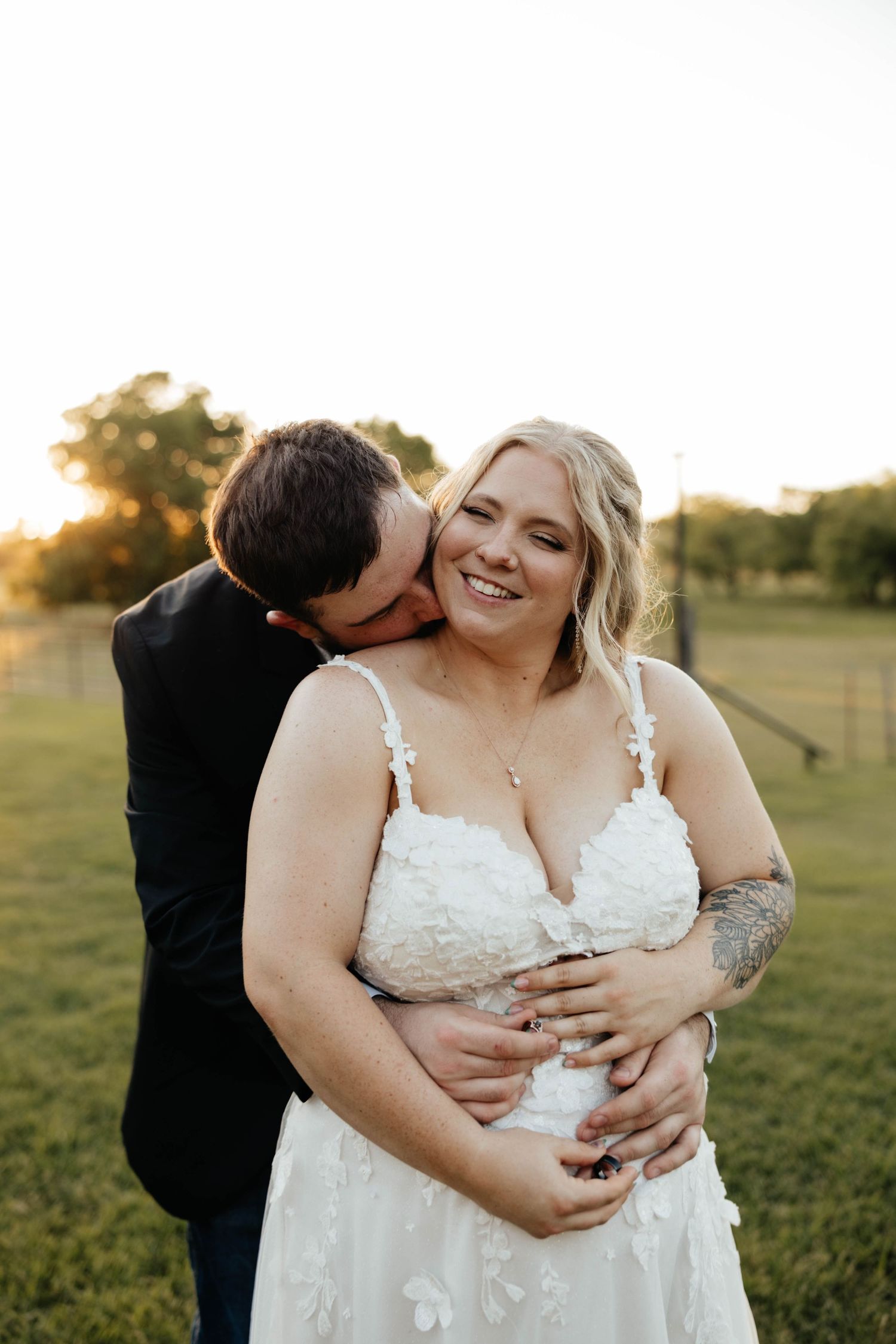 Groom kisses bride on cheek as she smiles in a floral wedding gown at golden hour.