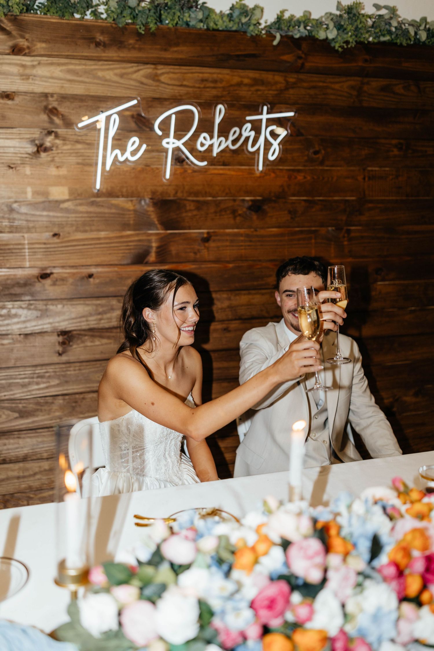 Newlyweds toast champagne beneath a neon sign reading The Roberts at their wedding reception.