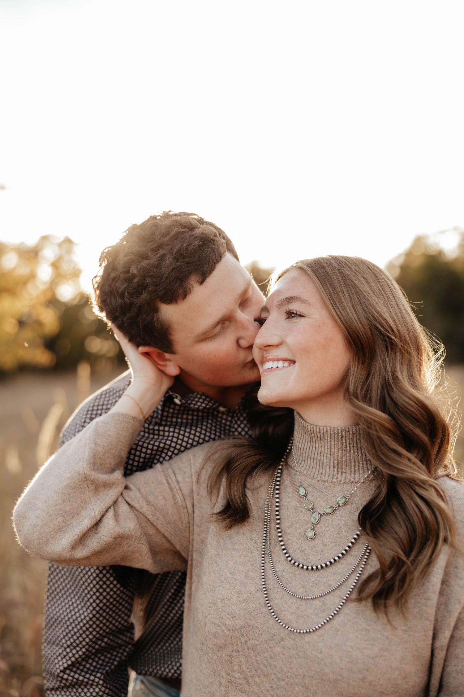 Man kisses smiling woman on cheek outdoors at golden hour, woman wearing layered turquoise necklaces.