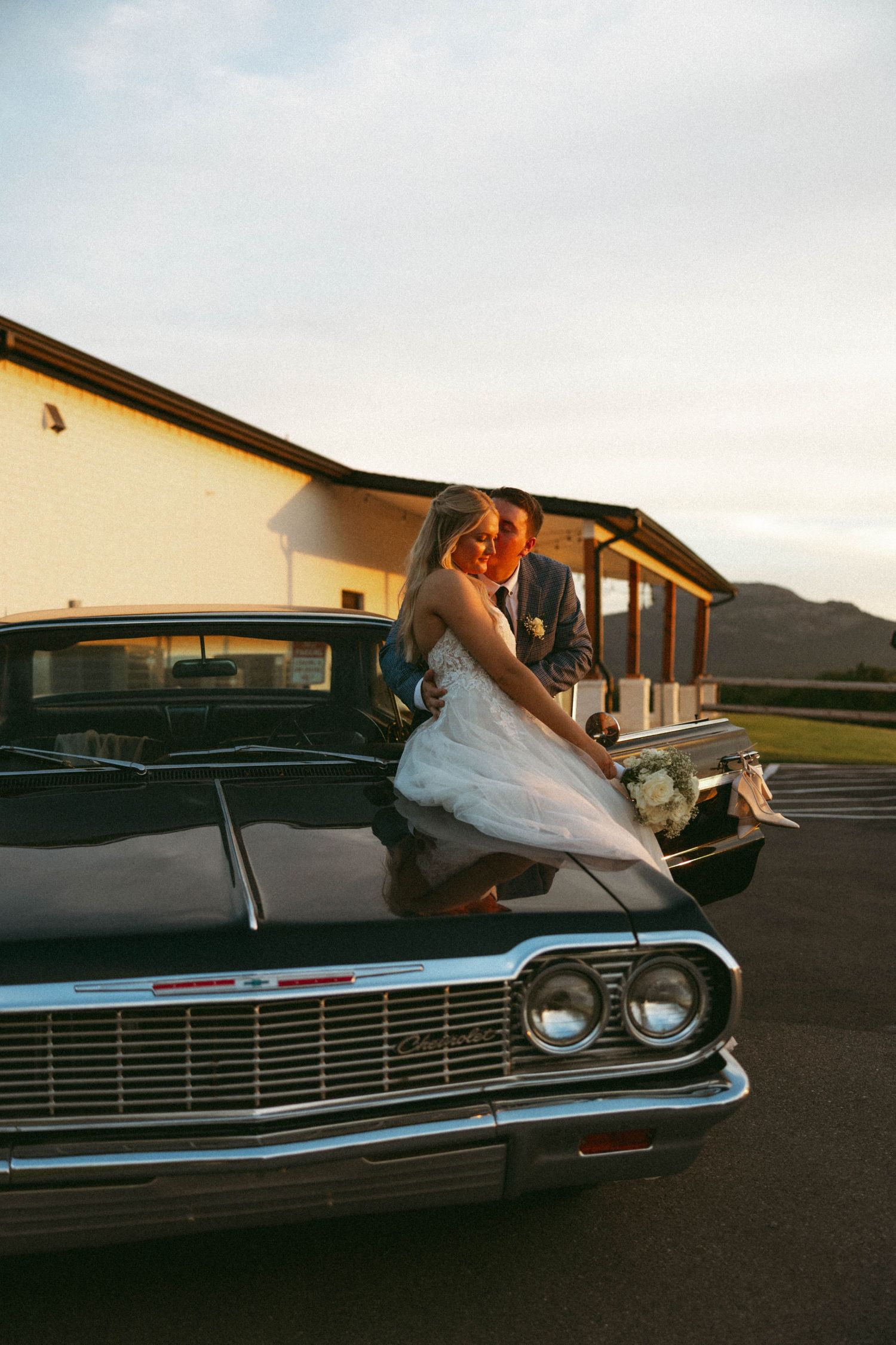 Bride and groom sharing a romantic moment on a classic vintage black car at golden hour sunset.