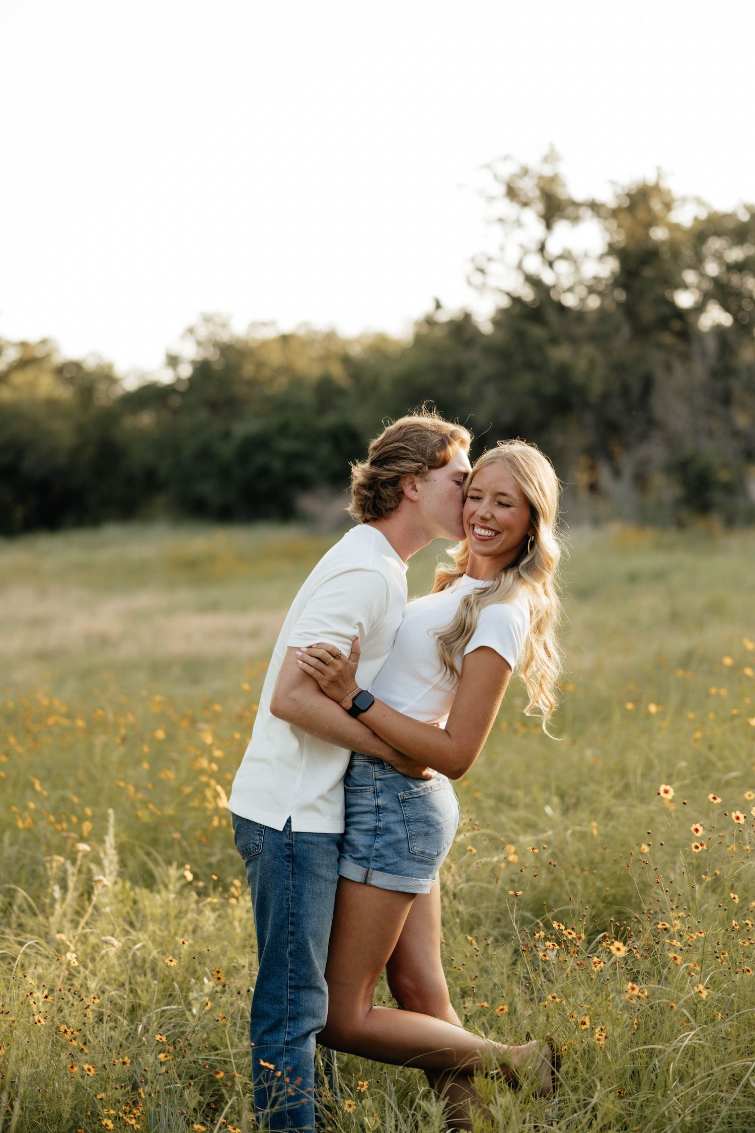 Couple embracing in a wildflower meadow at sunset, man kissing smiling woman in white tee and denim shorts.