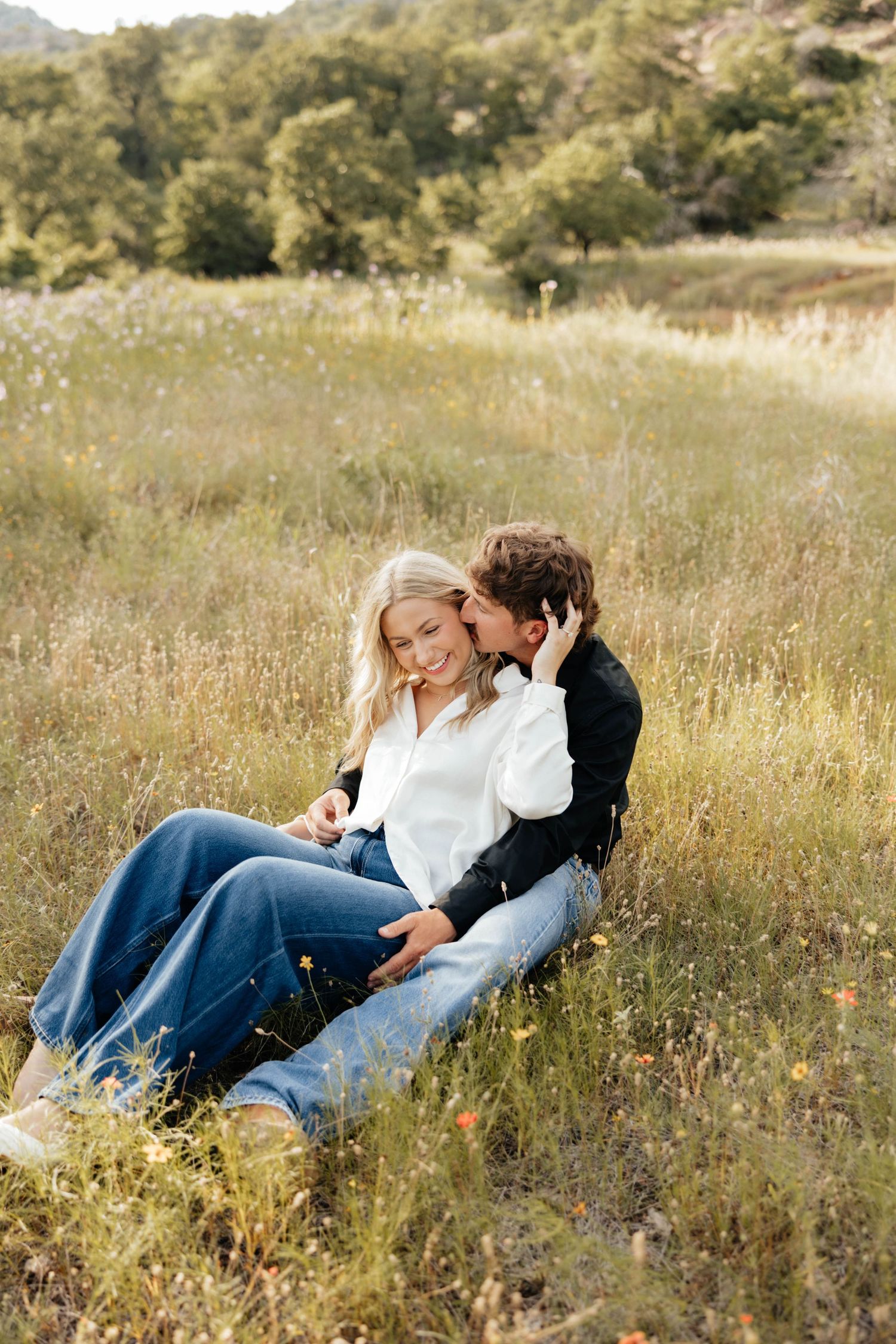 Couple sitting together in wildflower meadow, man whispering to smiling woman in white shirt.