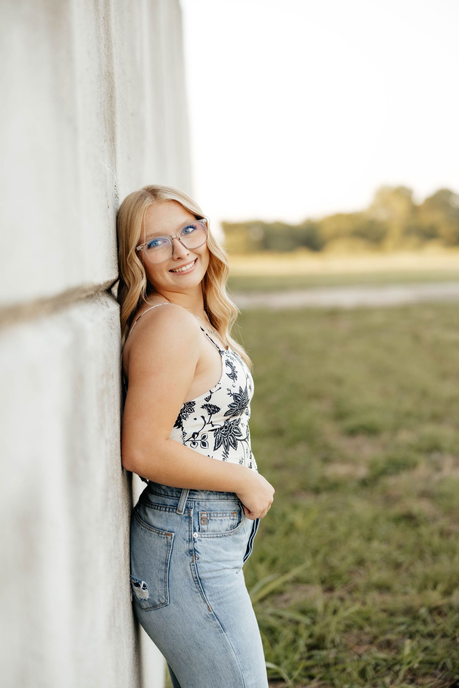 Smiling blonde woman with blue glasses leaning against white wall in floral top and jeans outdoors.