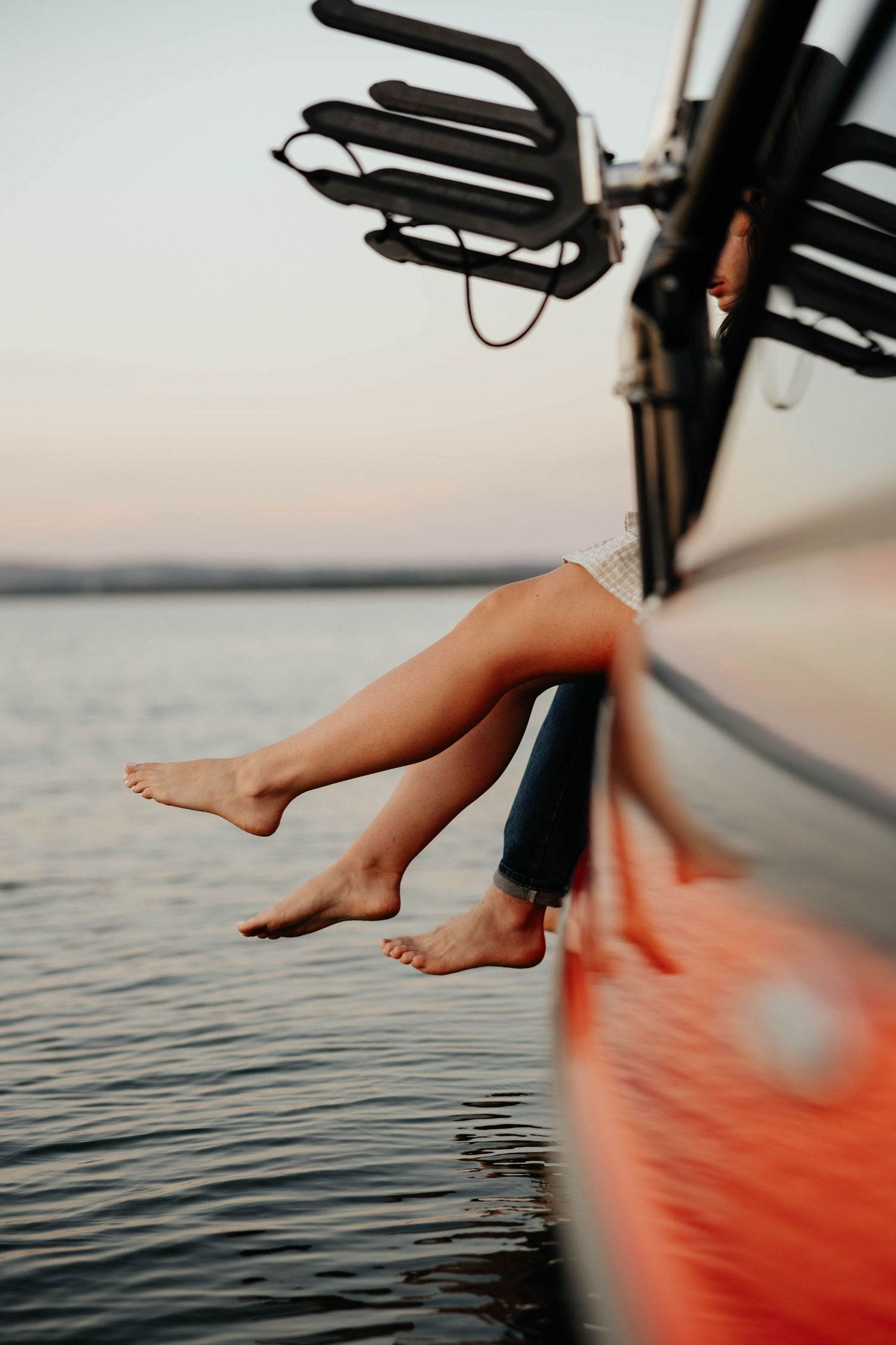 Bare legs dangling over the side of a red boat at sunset over calm water.