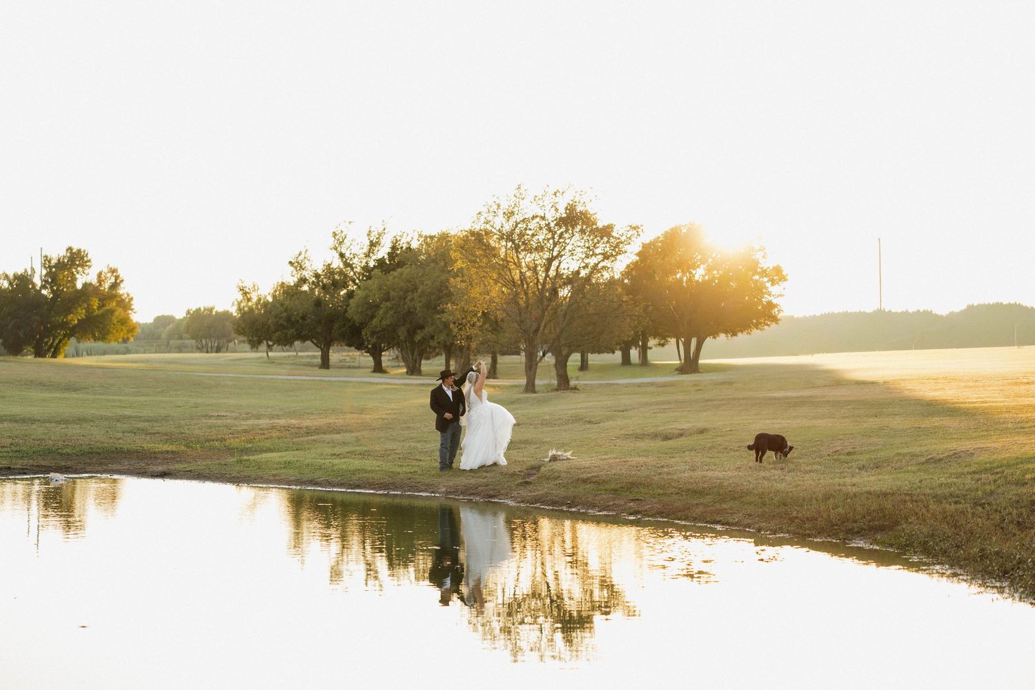 Bride and groom reflected in pond at golden hour surrounded by trees on golf course.