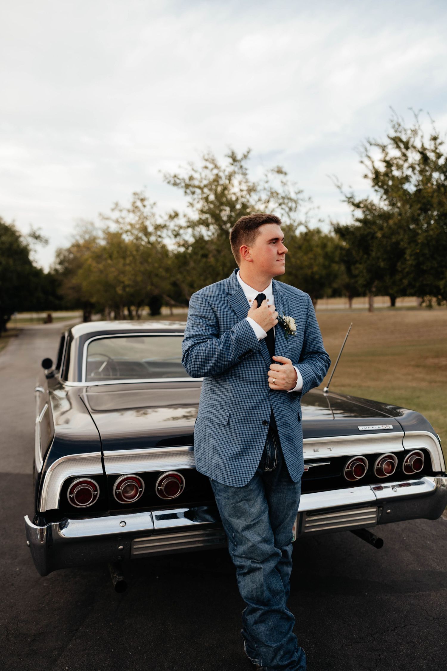 Groom in blue checkered blazer and jeans leaning against vintage black classic car in tree-lined setting.