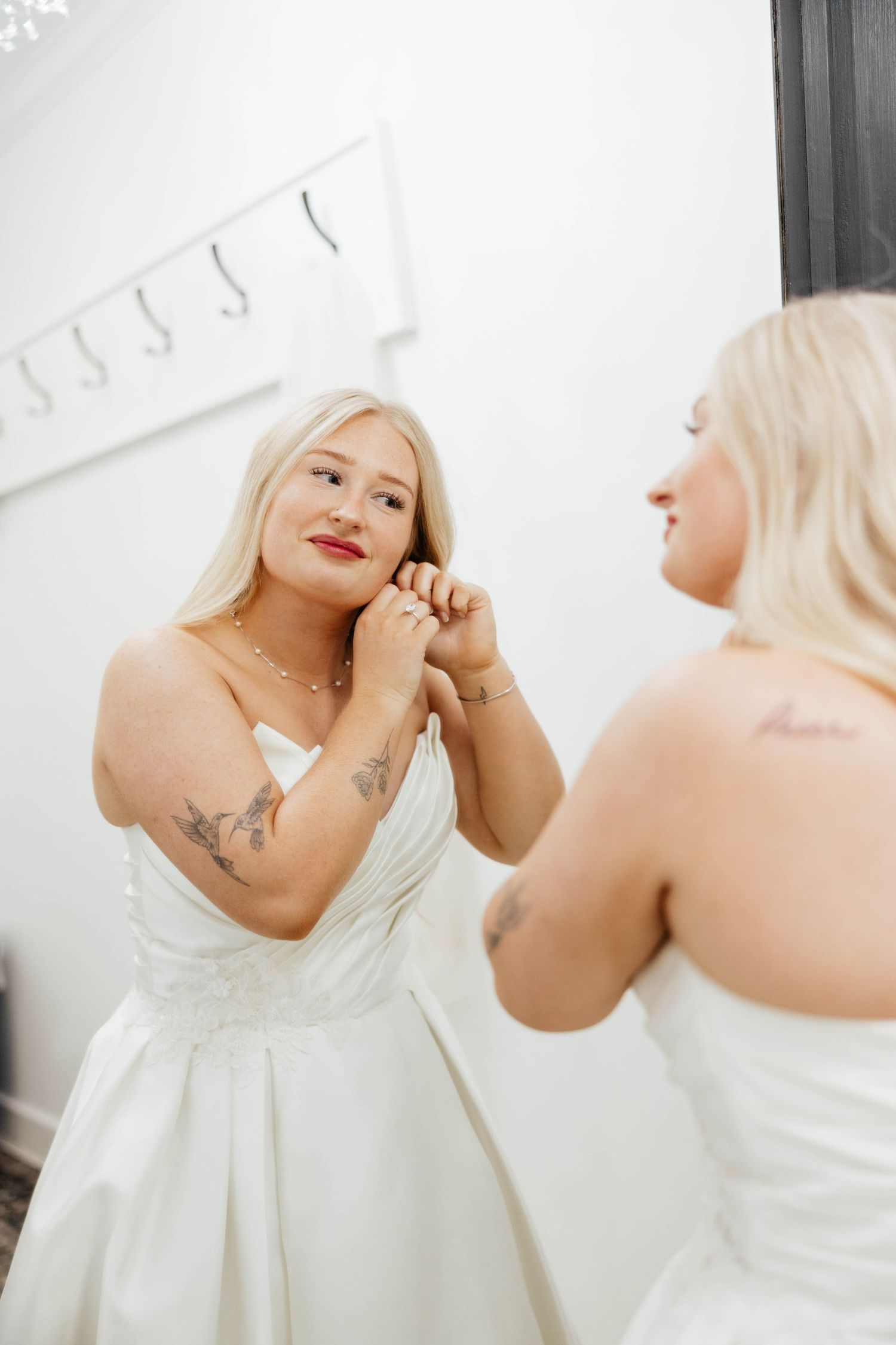 Blonde bride with tattoos adjusts her earring while getting ready in front of a mirror.