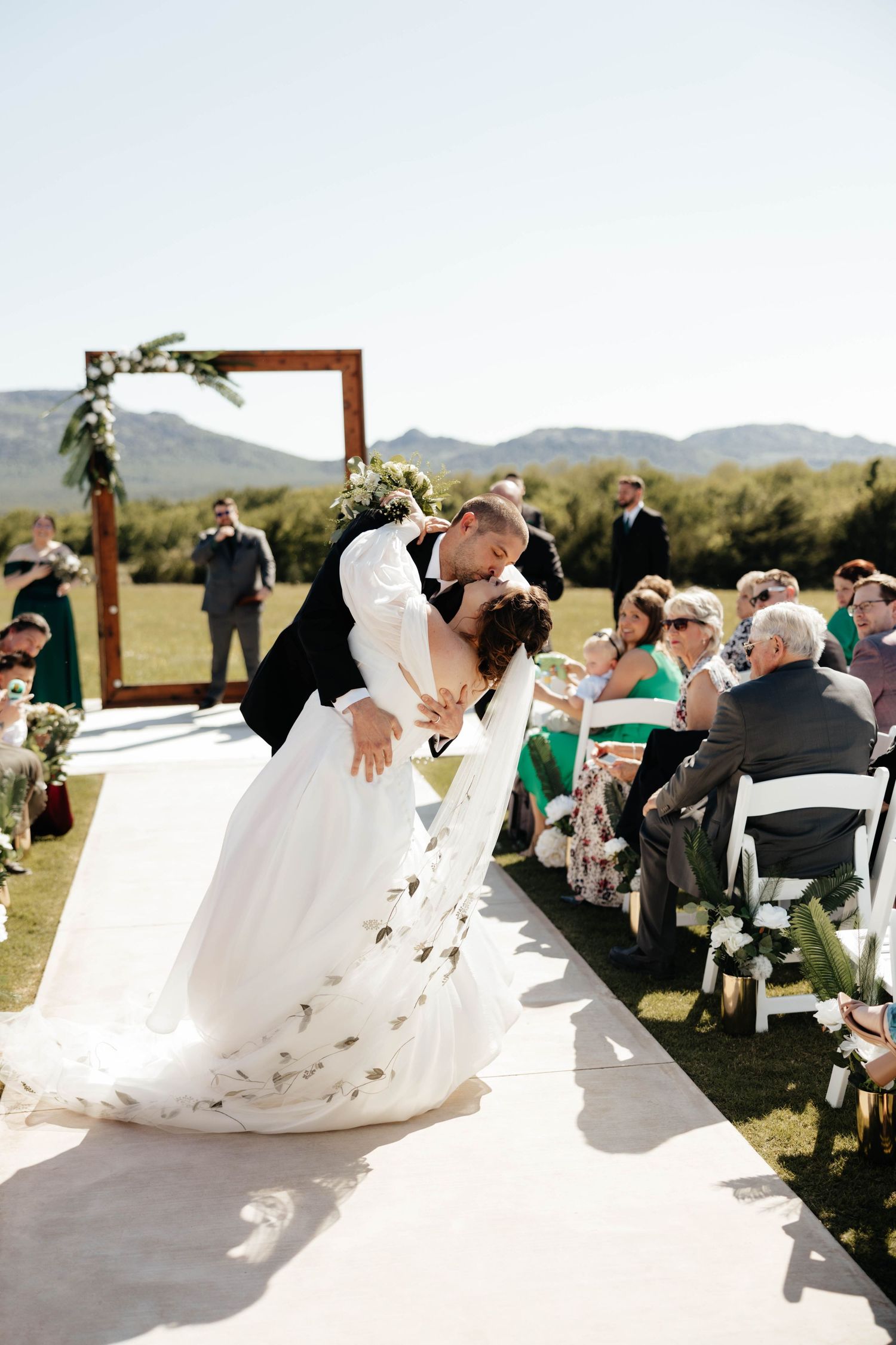 Groom dips bride for a kiss at outdoor wedding ceremony with mountain backdrop.
