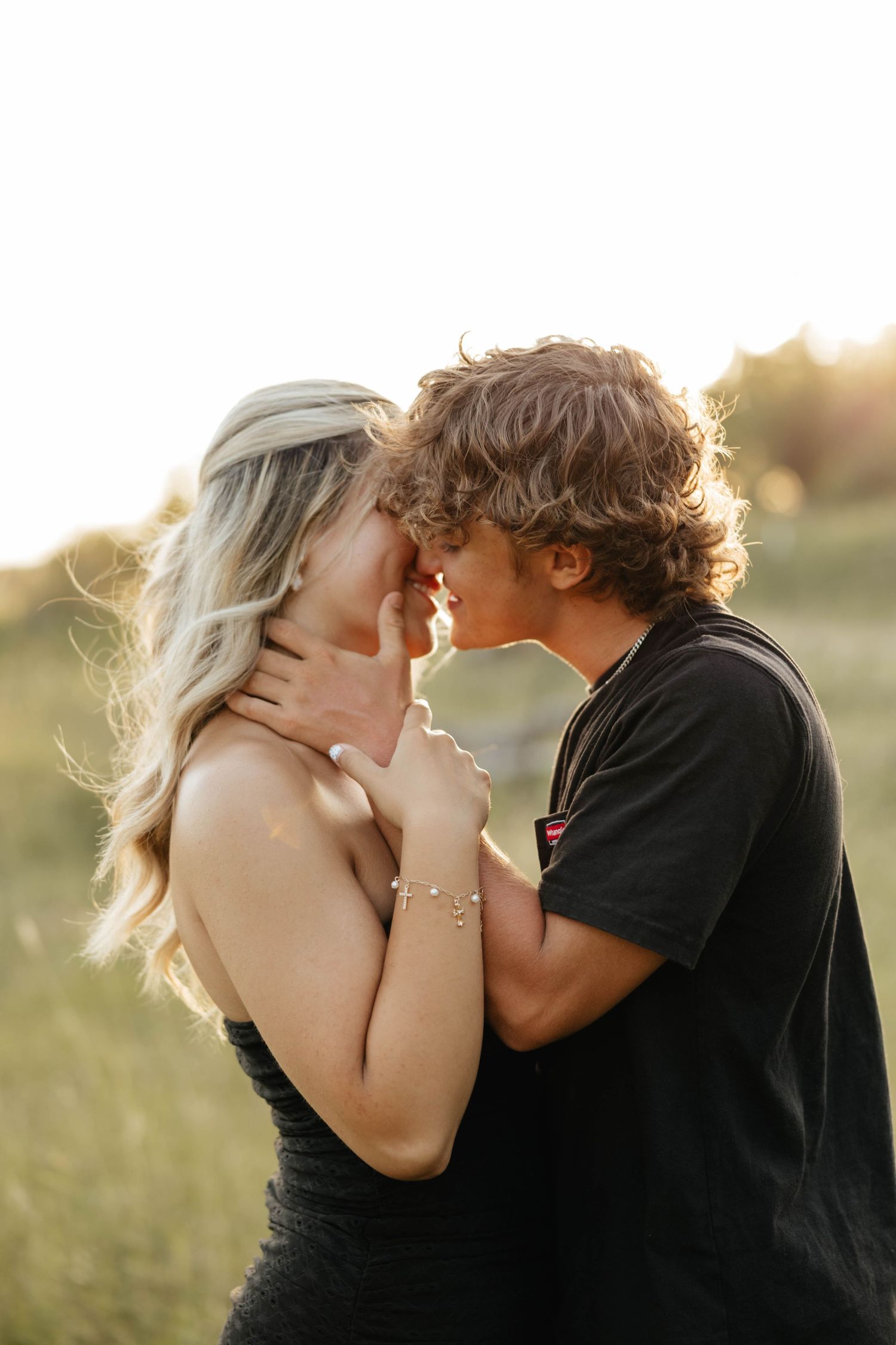 Couple sharing a romantic almost-kiss at golden hour in a sunlit field, woman in black dress, man in black tee.
