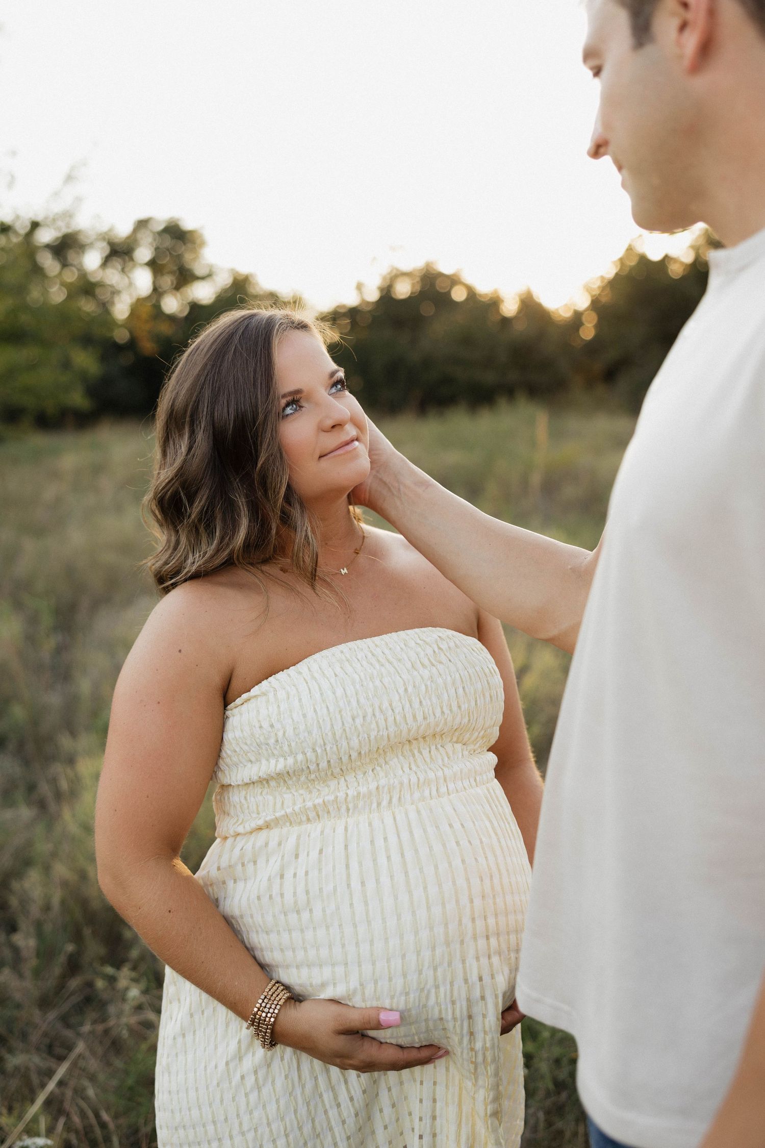 Pregnant woman in strapless cream dress gazes up at partner touching her face in sunlit field.