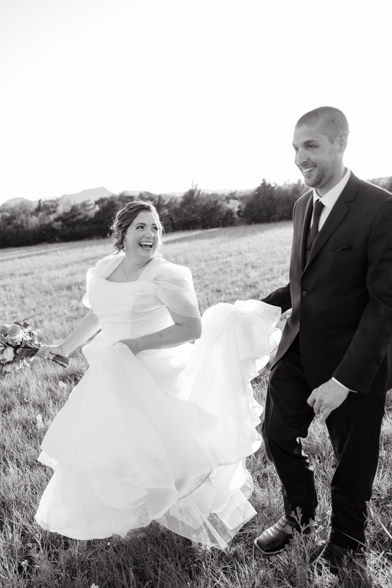 Black and white photo of laughing bride and groom walking through field on wedding day.