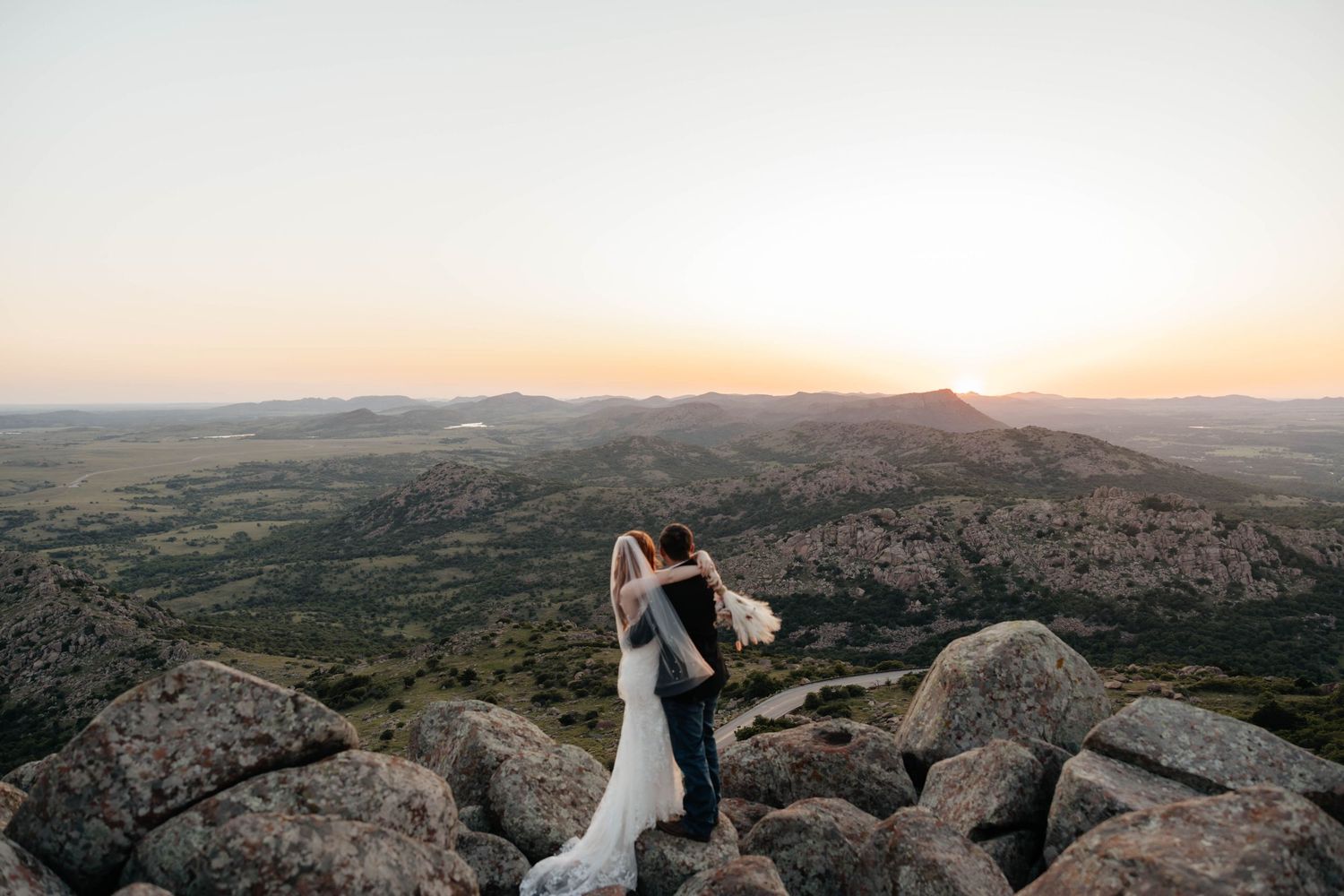Bride and groom embracing on a rocky mountaintop with a breathtaking panoramic sunset landscape.