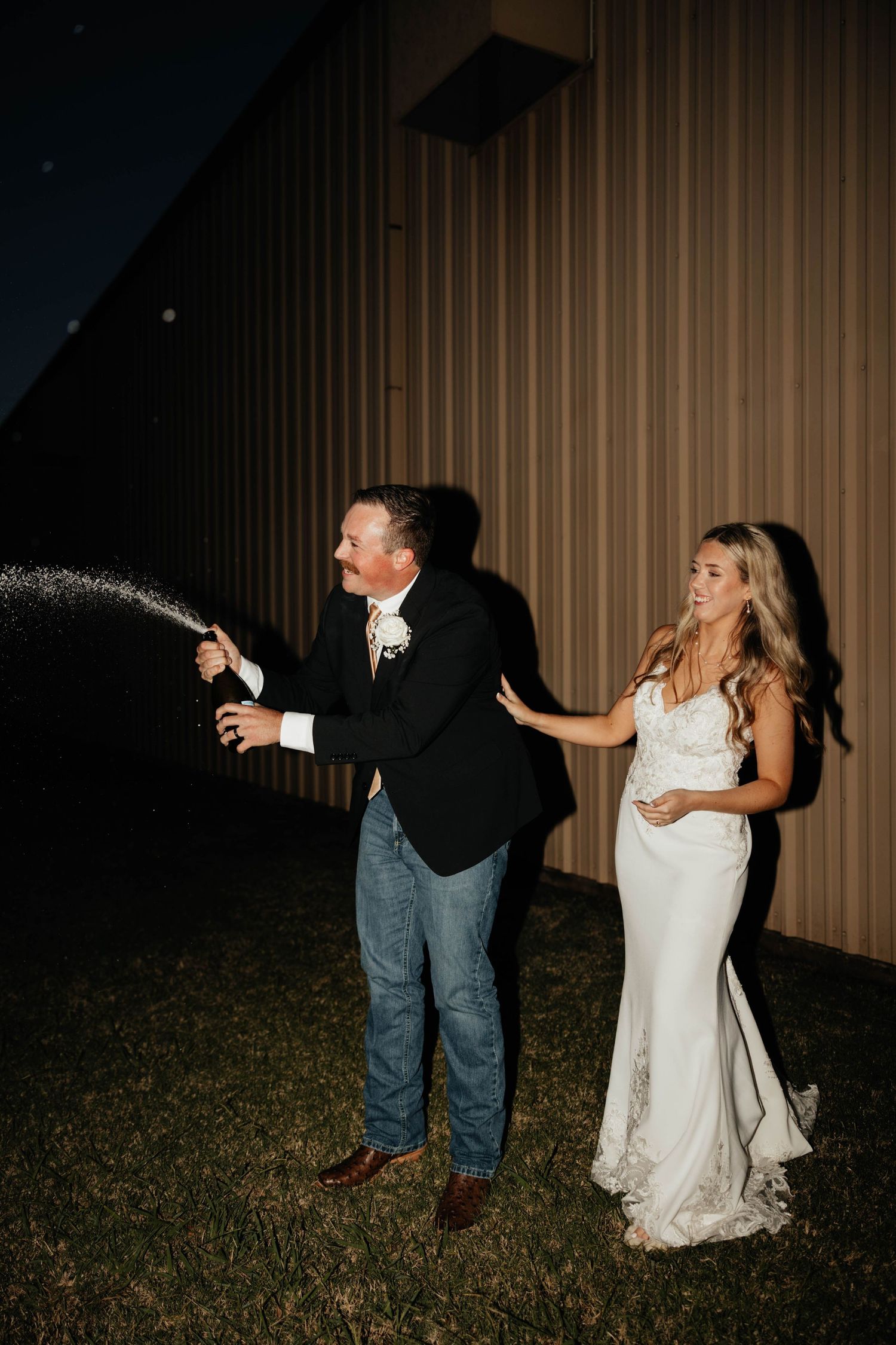 Groom popping champagne bottle outdoors at night while bride stands beside him smiling.