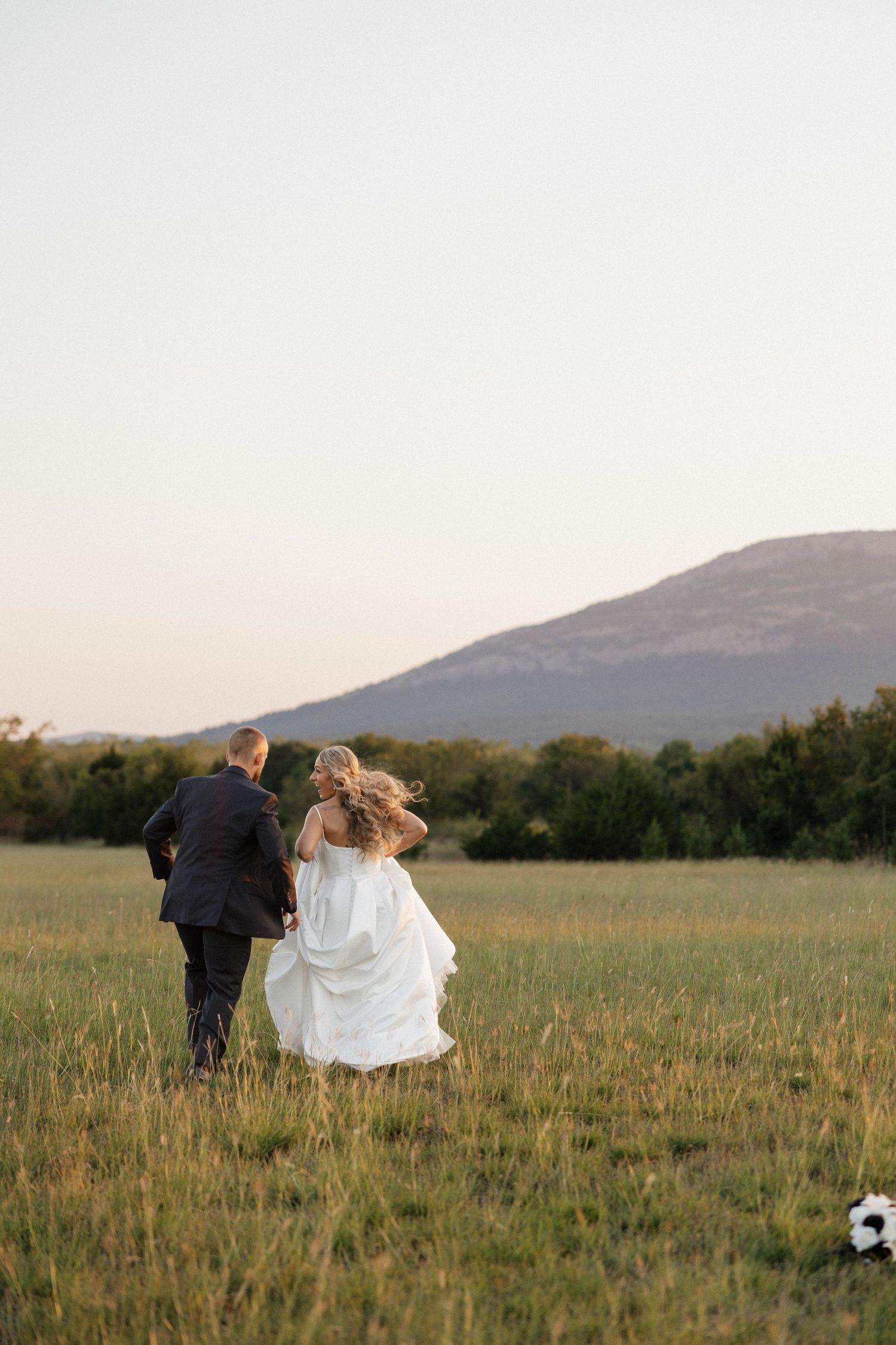 Bride and groom walk hand in hand through a green meadow with a mountain backdrop at dusk.