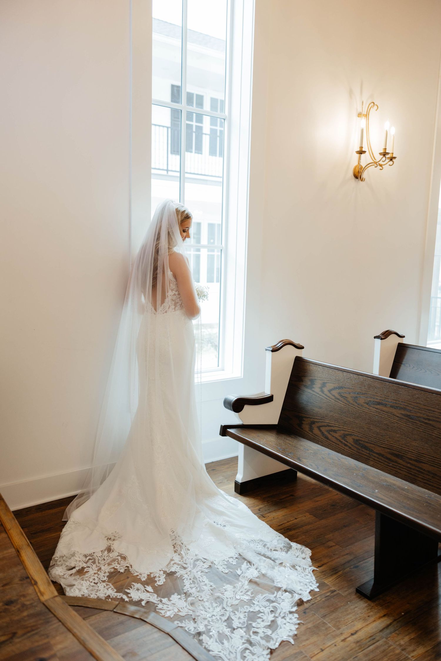 Bride in lace wedding gown with cathedral veil gazing out tall chapel window beside wooden pew.