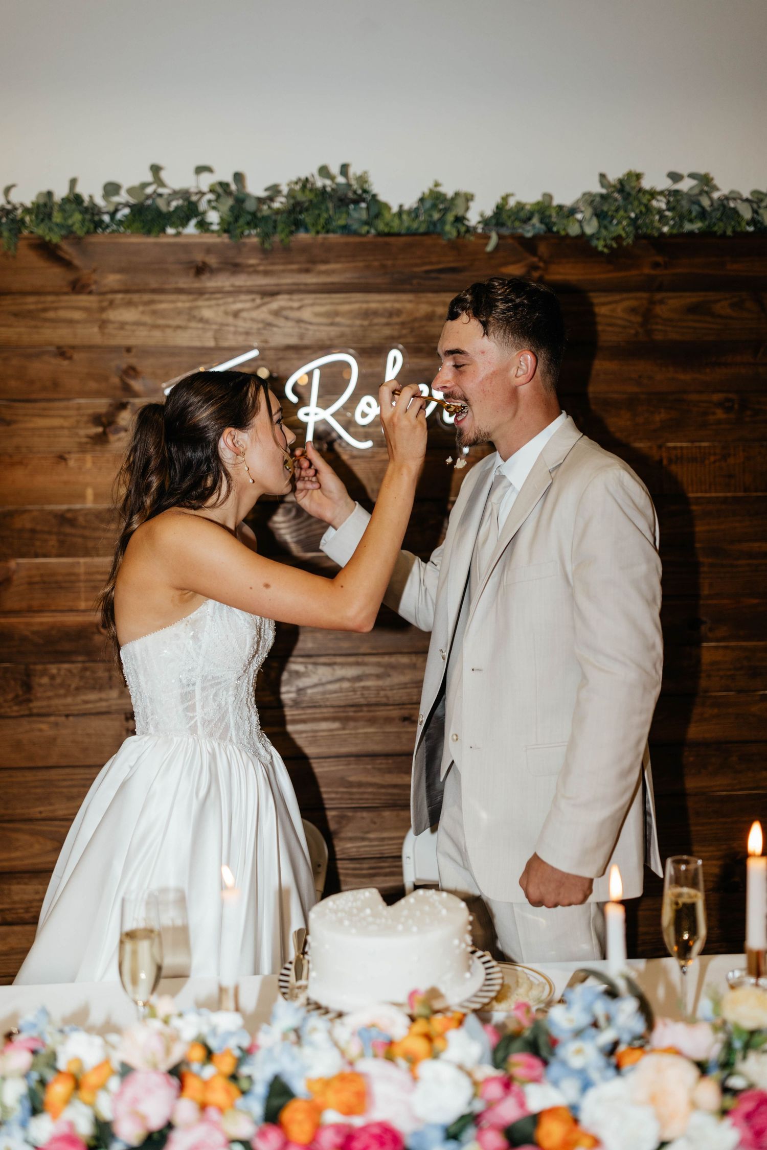 Bride feeds groom a piece of wedding cake in front of a glowing neon sign backdrop.