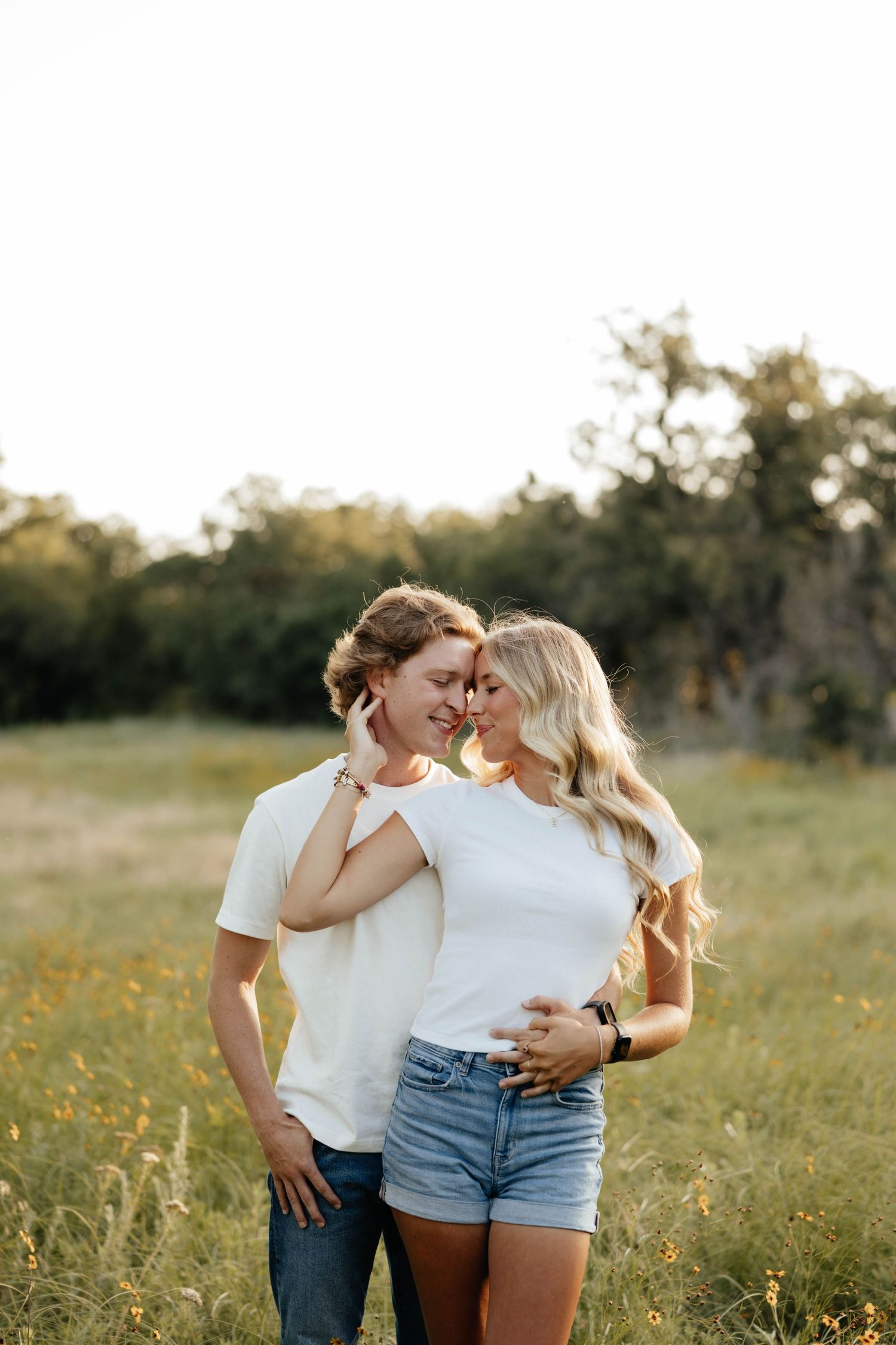 Romantic couple in matching white tees share an intimate moment in a sunlit green meadow at golden hour.
