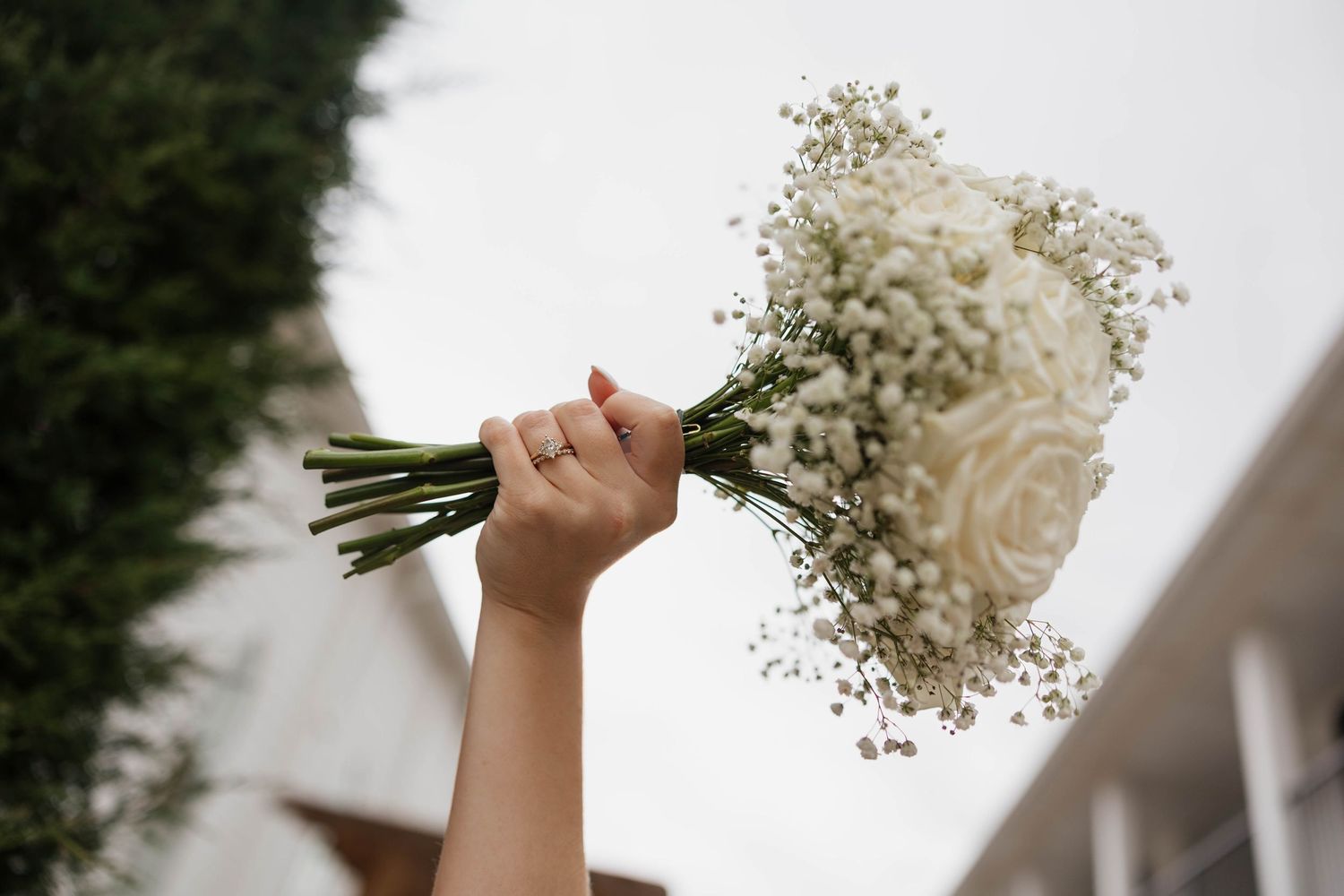 Hand holding a white rose and baby's breath bridal bouquet raised against a cloudy sky.