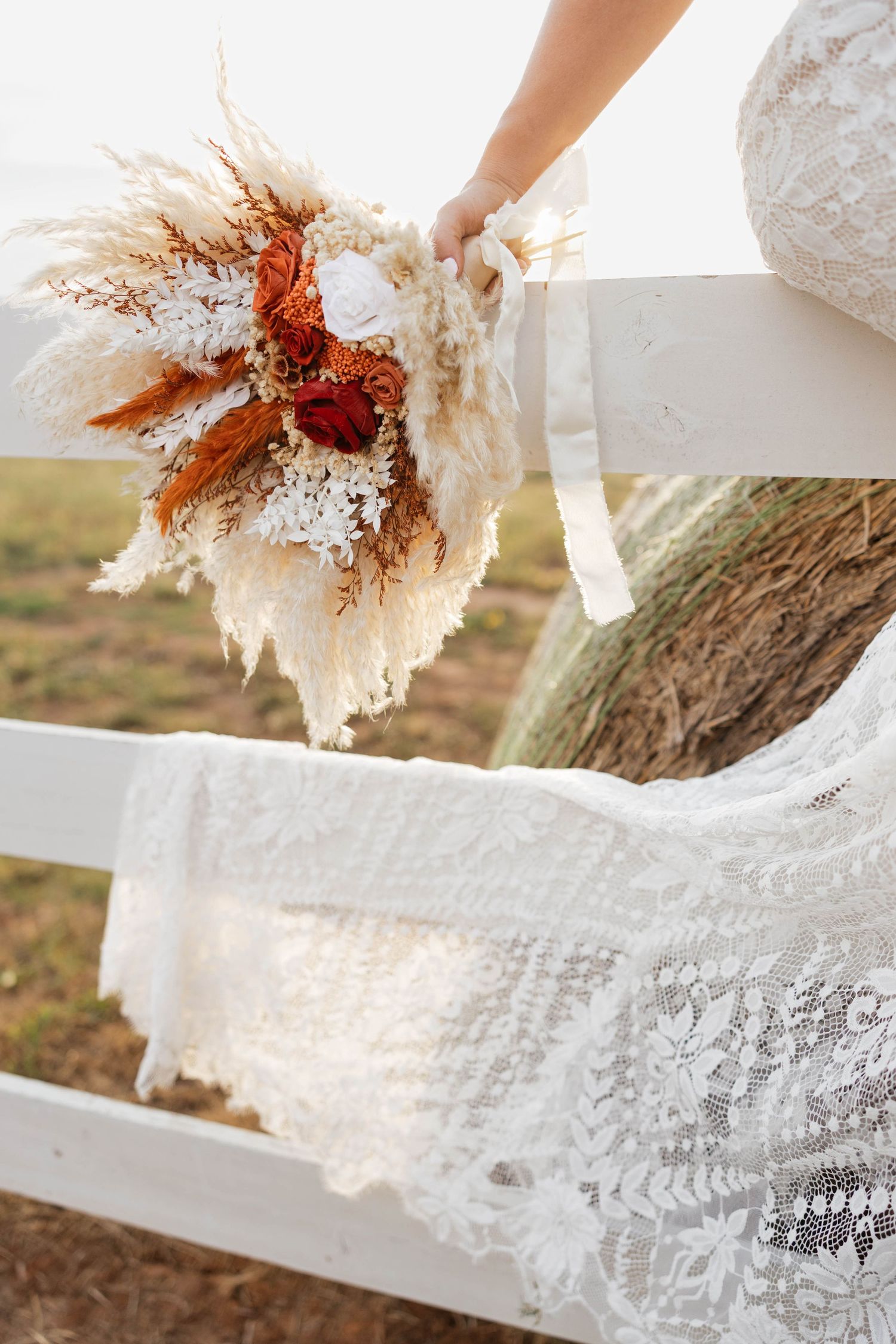 Bride holding a boho dried floral bouquet with pampas grass near a white fence.